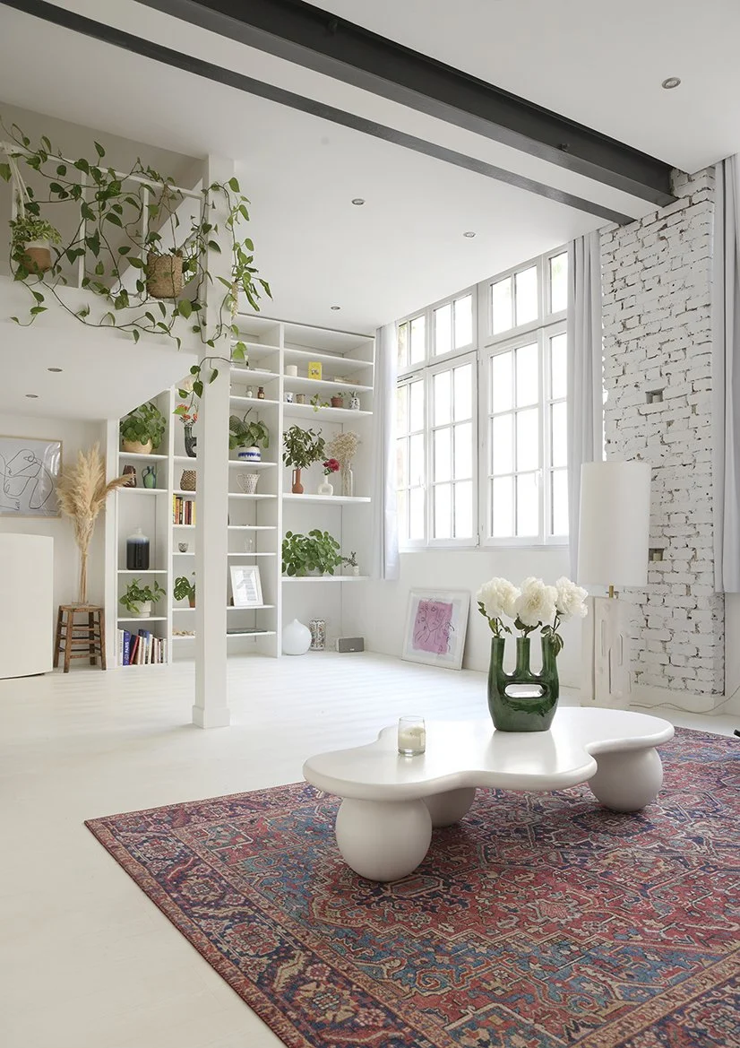 Salon lumineux au style moderne avec canapé blanc, table basse organique, tapis persan, plantes vertes et mur en briques blanches.
