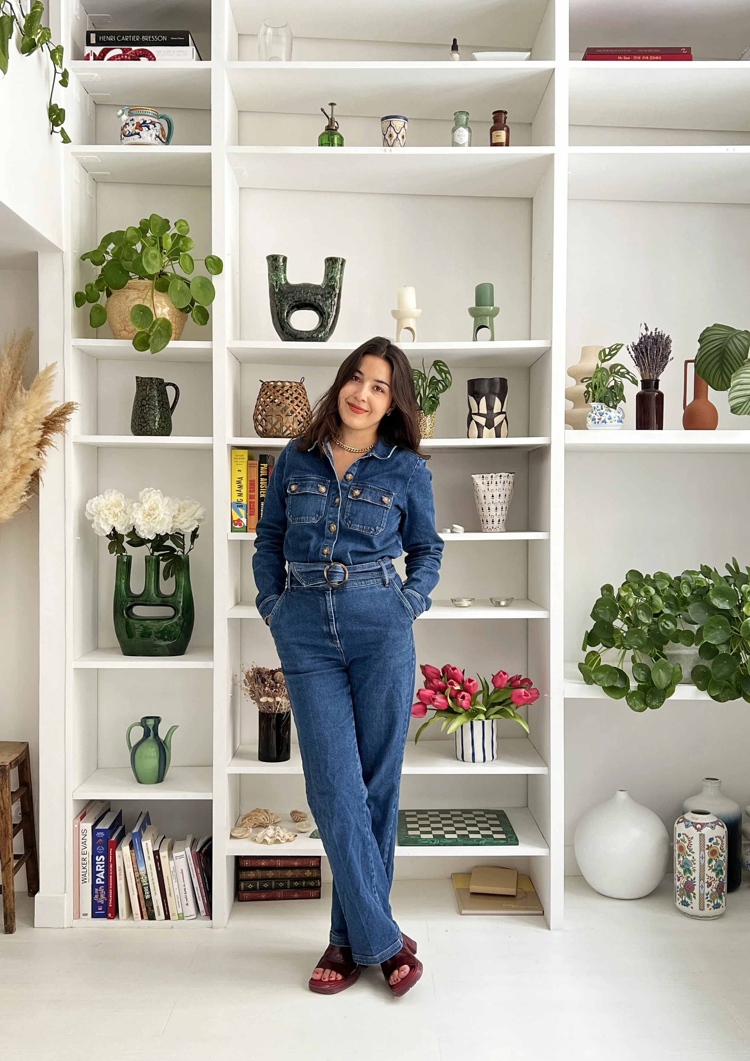Femme en combinaison en jean posant devant une bibliothèque blanche décorée de plantes, vases, livres et fleurs.