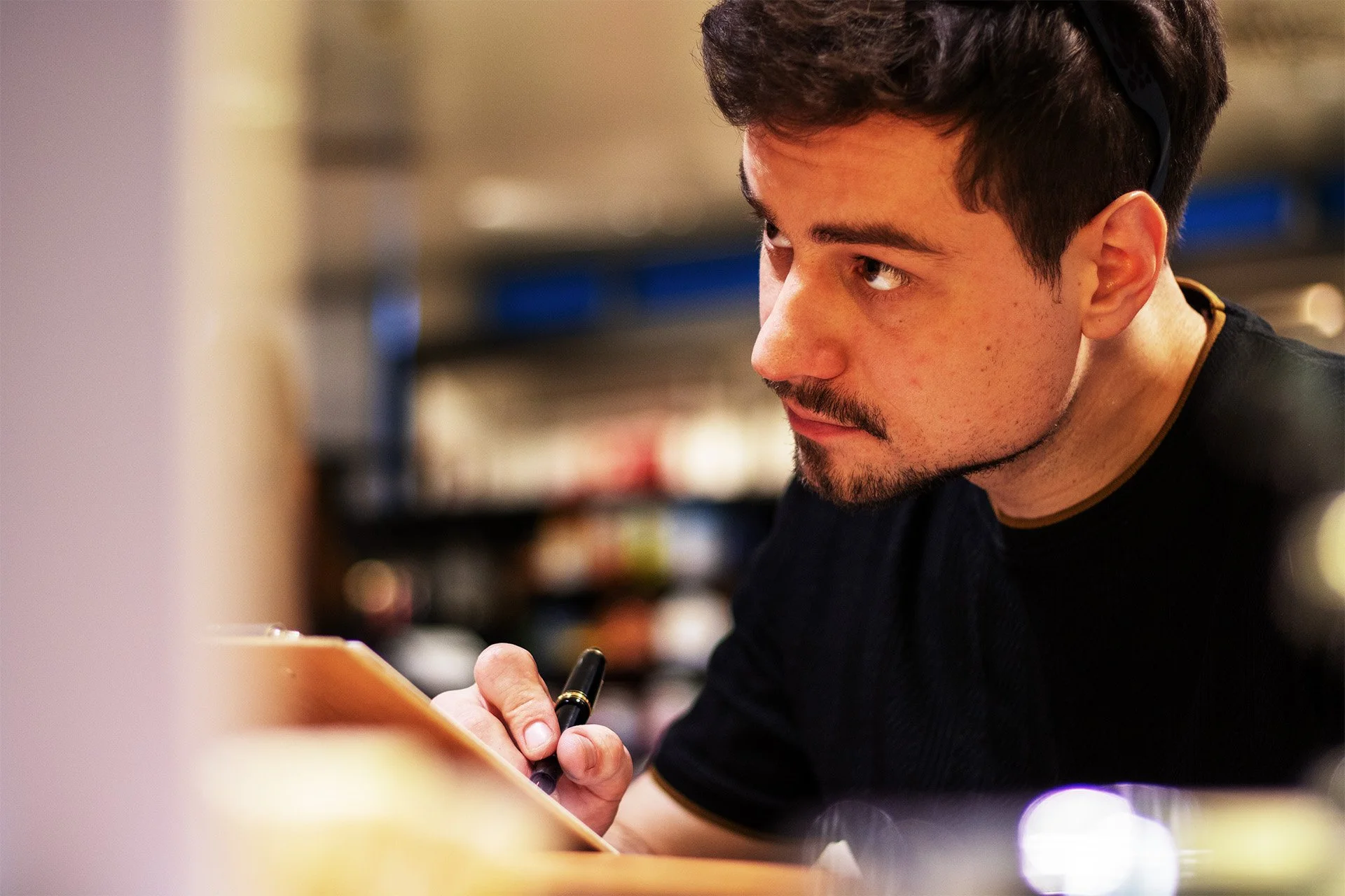 Junger Mann mit dunklen Haaren, Schnurrbart und Bart schreibt mit einem schwarzen Stift auf eine orangefarbene Notizblock im Supermarkt, im Hintergrund Regale mit Produkten, die unscharf sind.