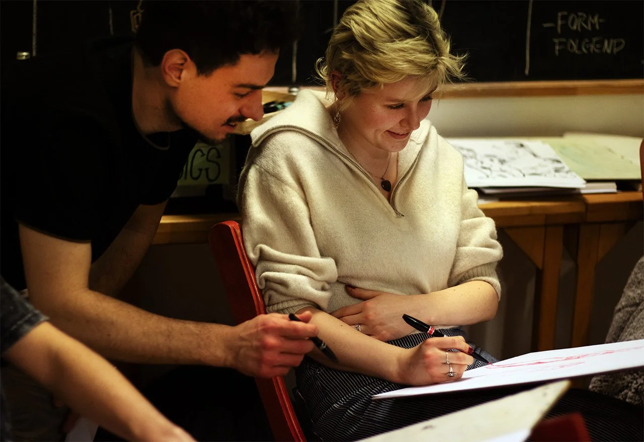 Zwei Menschen, ein Mann und eine Frau, sitzen an einem Tisch, die Frau schreibt auf einem großen Blatt Papier, während der Mann ihr schreibt. Sie lächeln und scheinen an einem gemeinsamen Projekt zu arbeiten.