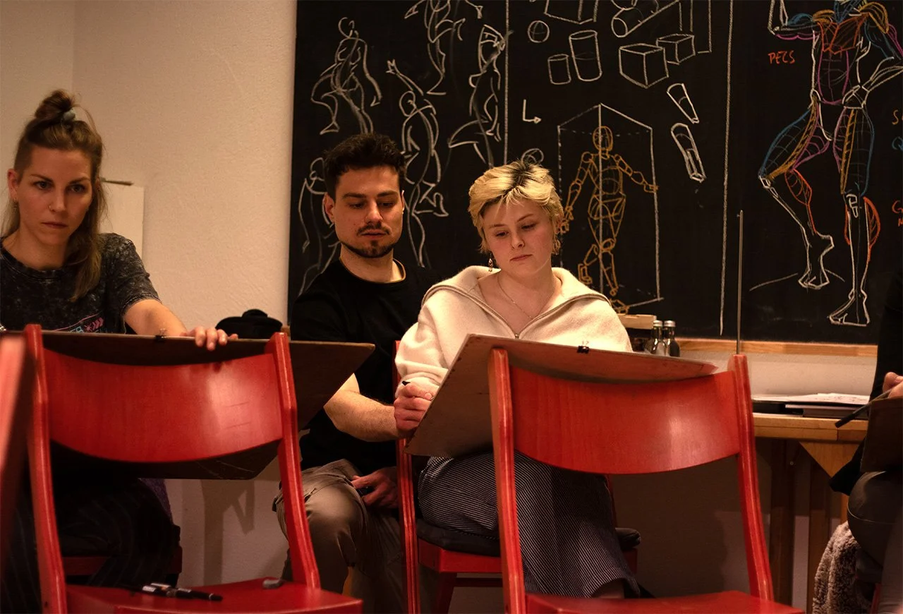 Drei junge Erwachsene sitzen an Tischen in einem Raum, in dem eine Tafel mit chemischen Anatomie-Skizzen im Hintergrund ist.