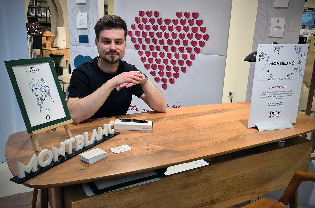 Mann sitzt an einem Holztisch bei einer Montblanc Veranstaltung, im Hintergrund eine Wand mit Herzdekoration, rechts ein Whiteboard mit Informationen über ein Live-Portrait-Event.