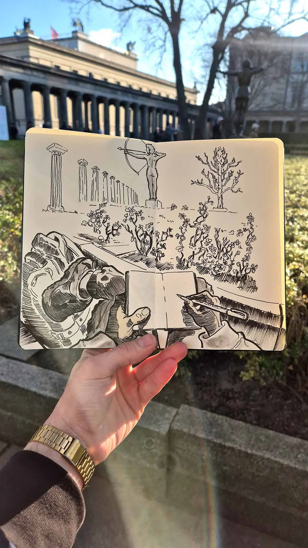 Eine Hand hält ein Skizzenbuch mit einer handgezeichneten Illustration vor einer Parklandschaft mit einer Statue, Bäumen und monumentalen Säulen im Hintergrund. Das Skizzenbuch zeigt eine Skizze eines Fahrrads, das auf einer Straße zeichnet, während die andere Hand mit einem Stift zeichnet.