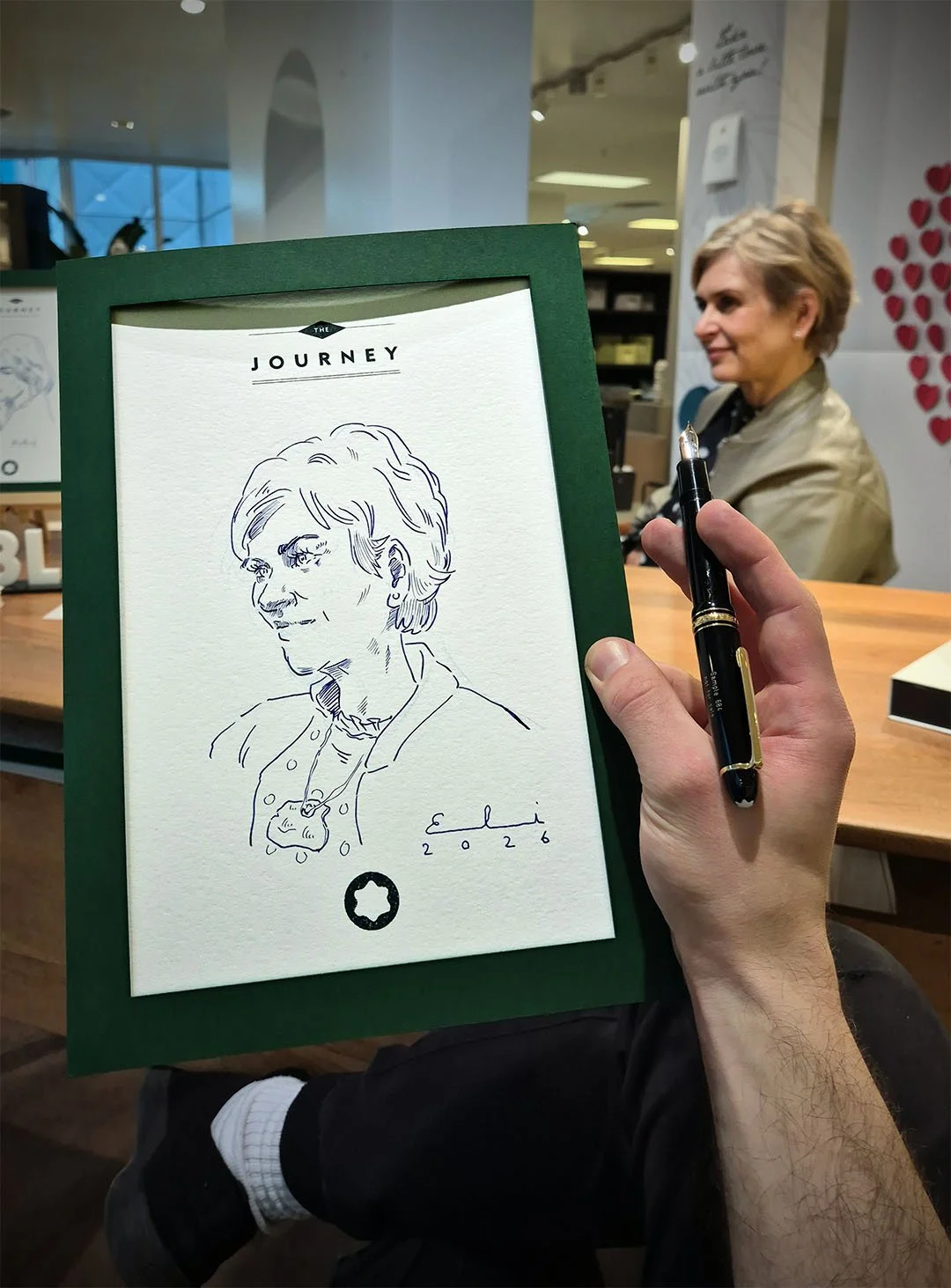 Zeichnung eines lächelnden Mannes auf einem weißen Papier in einer grünen Mappe, die an einem Tisch gehalten wird, mit einem Signatur- und Datumsvermerk darunter. Im Hintergrund eine Frau sitzt und spricht in einem gut beleuchteten Raum.