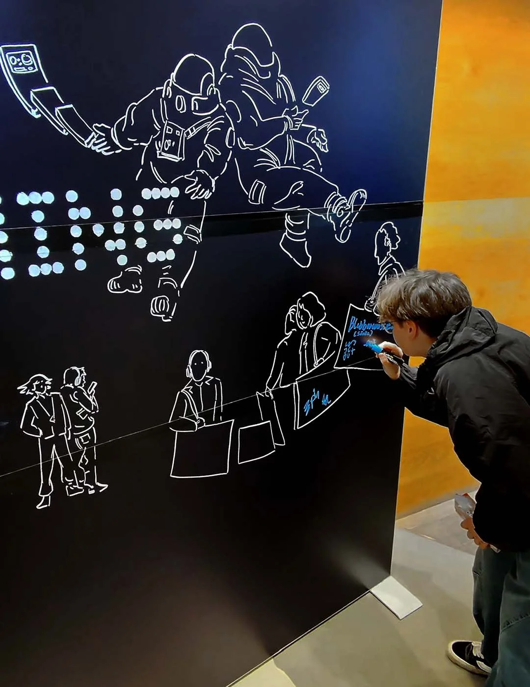 Ein junger Mann schreibt mit einem blauen Marker auf eine schwarze Wand mit weißen, linienartigen Illustrationen von Menschen, die in einer Warteschlange an einem Informationsschalter stehen, während im Hintergrund geometrische Flächen in Gelb- und O
