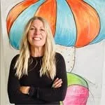 A woman with long blonde hair smiling with arms crossed in front of a colorful mural of oversized mushrooms.