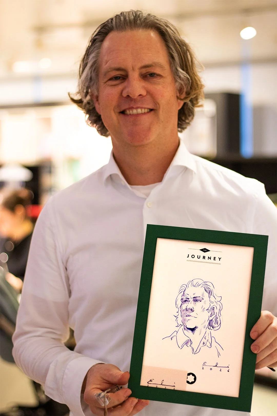 Ein Mann in weißem Hemd hält ein gerahmtes Bild mit der Überschrift "The Journey" und einer handskizzierten Porträtzeichnung. Er lächelt in die Kamera.