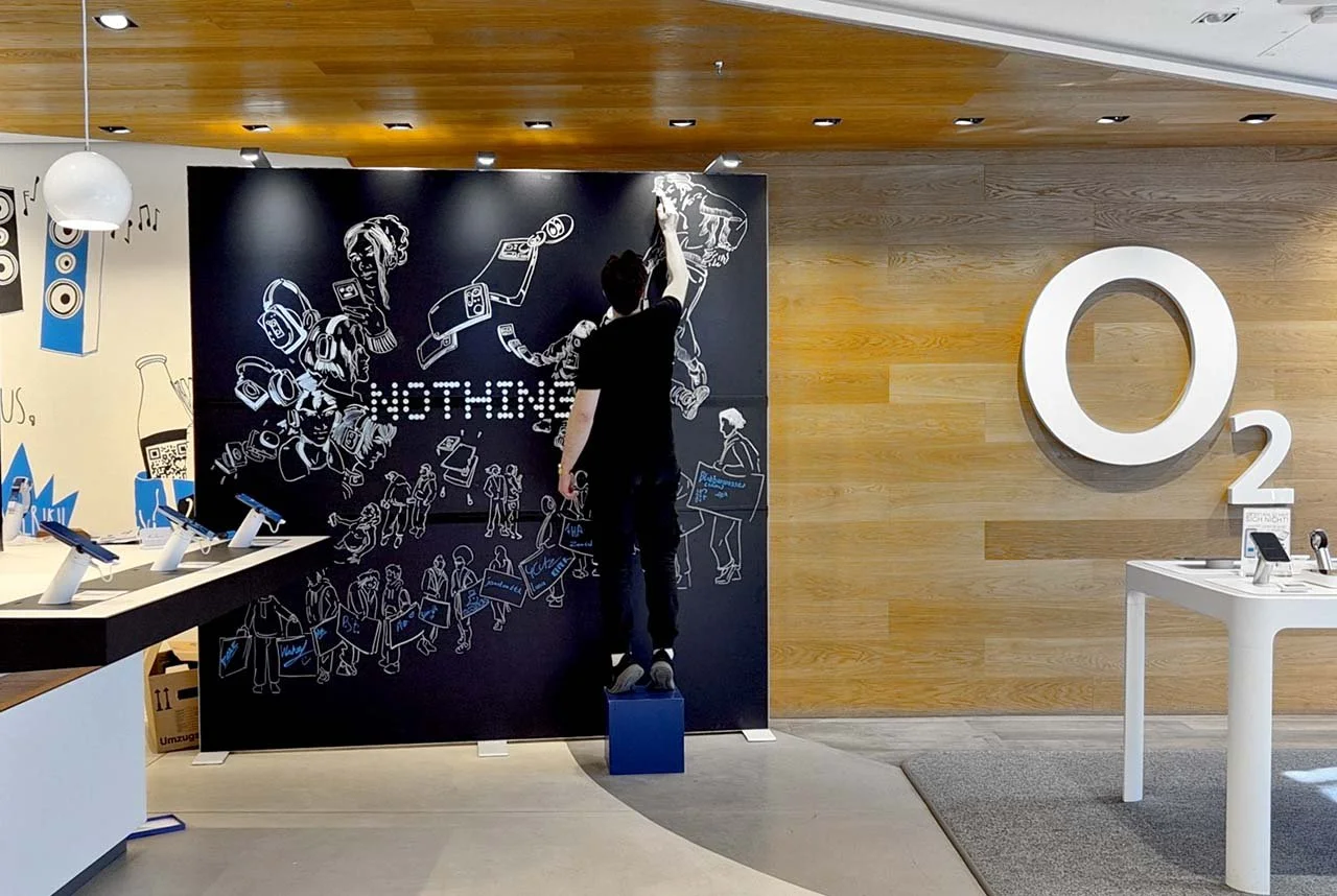 Mensch, der an einer Tafel mit der Aufschrift 'NOTHING' malt, in einem Laden, im Hintergrund ein großes O2-Logo an einer Holzwand und eine Tafel mit weißen Linien, das Ambiente ist modern eingerichtet.