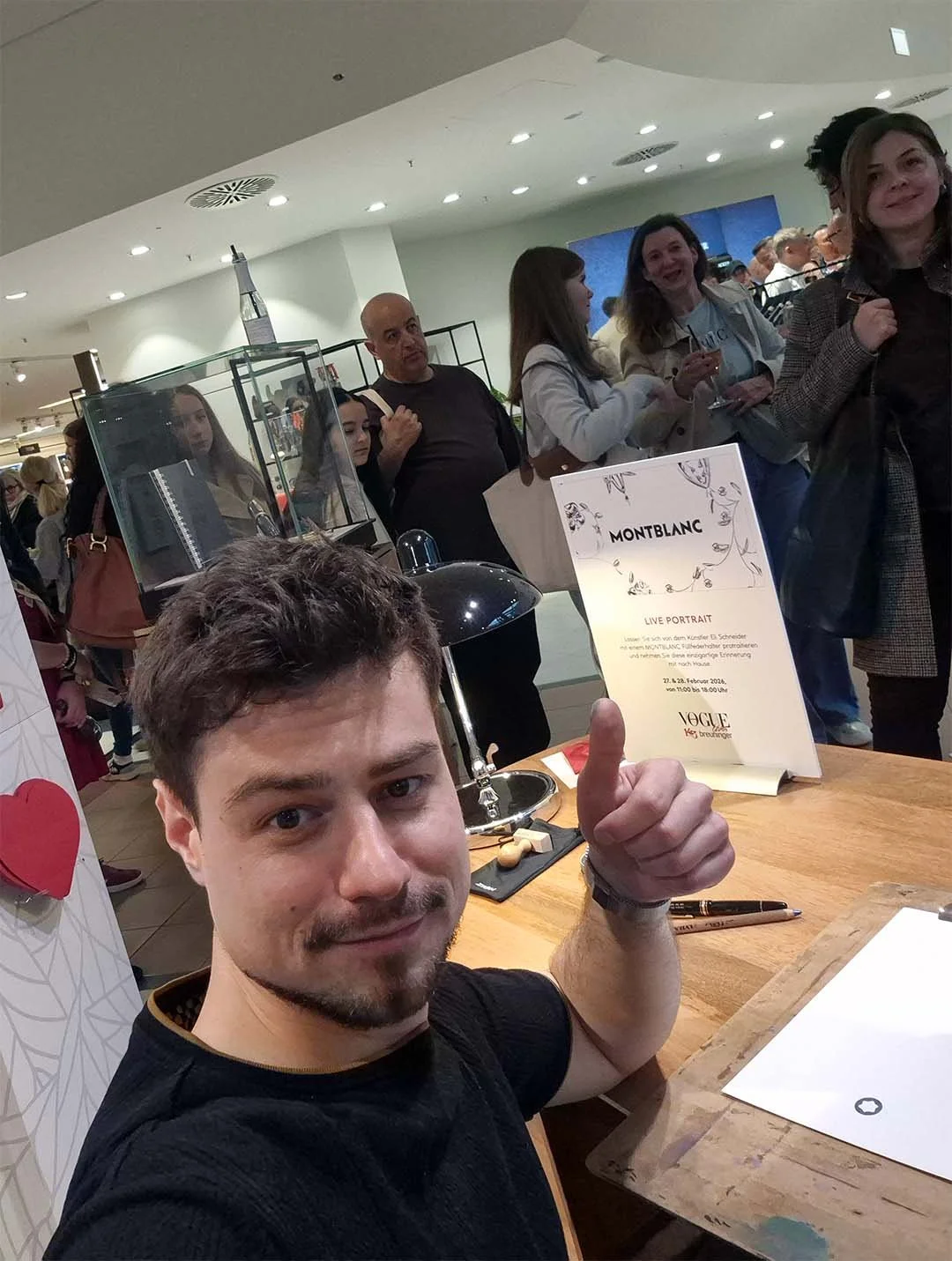 Junge Mann in Schwarz gibt Daumen hoch vor einem Holztisch, im Hintergrund eine Menschenmenge in einem Laden oder Ausstellungsraum, mit einem Montblanc-Poster.