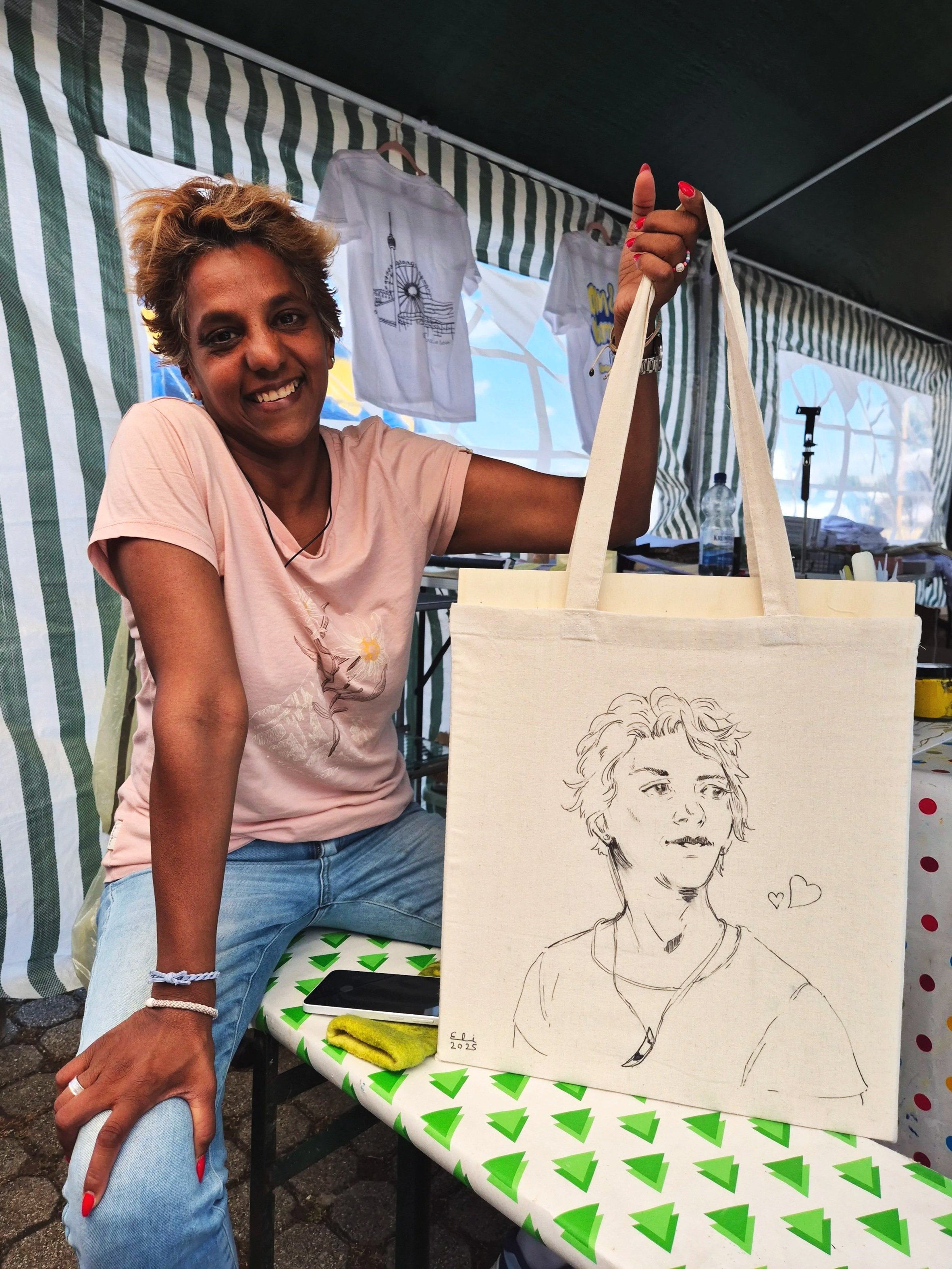 A woman with short, curly hair smiling at the camera while holding up a tote bag with a line drawing of a woman's face on it. She is sitting at a table with a green and white patterned tablecloth, underneath a striped canopy. Behind her, there are t-shirts hanging on a line and a water bottle.