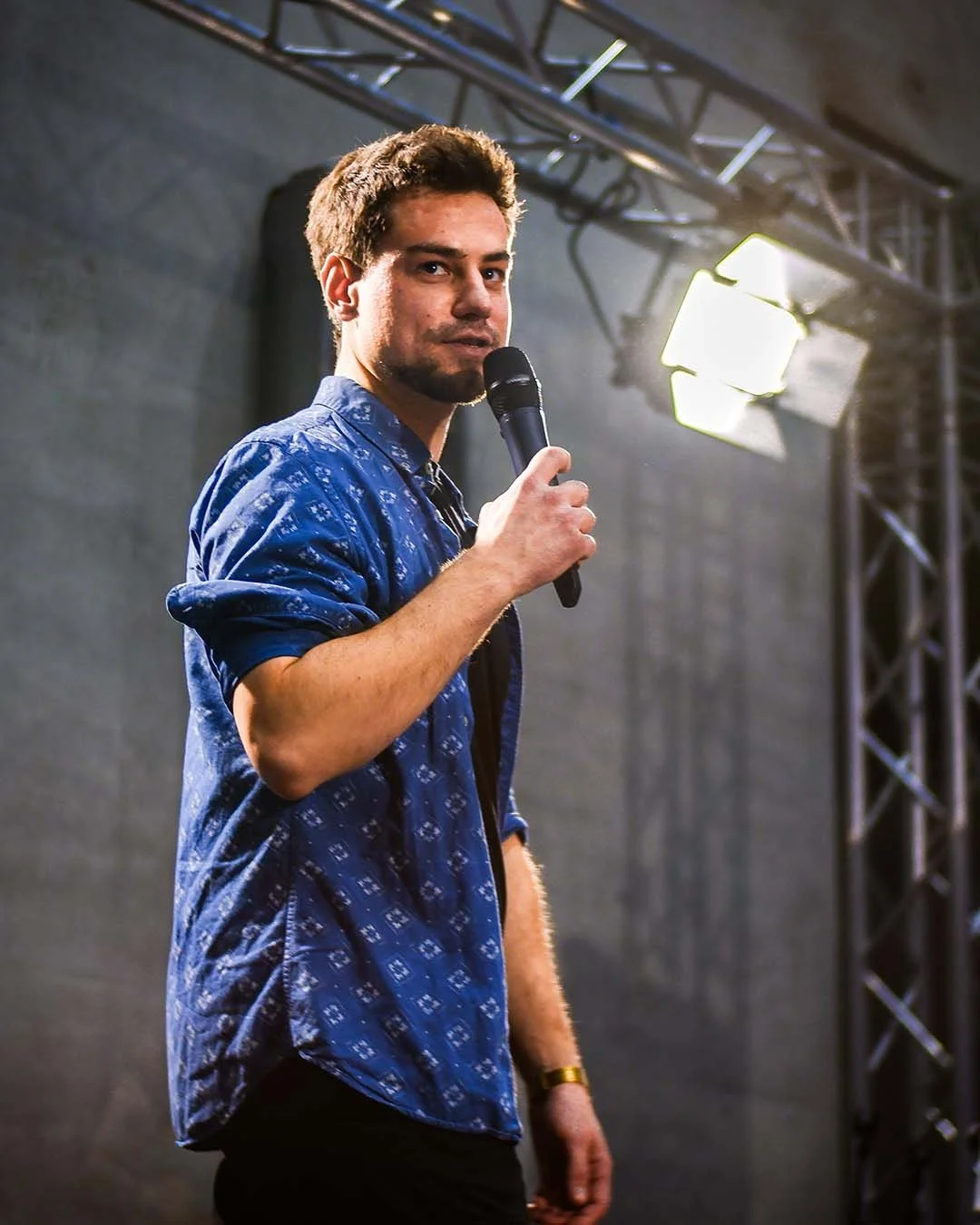 Mann hält Mikrofon und spricht vor Publikum. Hintergrund mit Bühnenbeleuchtung und Bühnenaufbau.