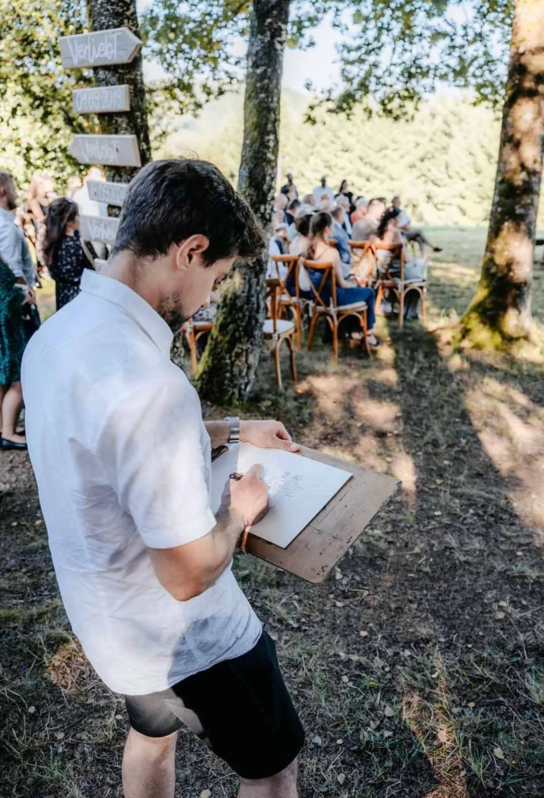 Ein Mann im weißen Hemd schreibt bei einer Outdoor-Hochzeit in einem Wald. Im Hintergrund sitzen Gäste auf Stühlen, umgeben von Bäumen.