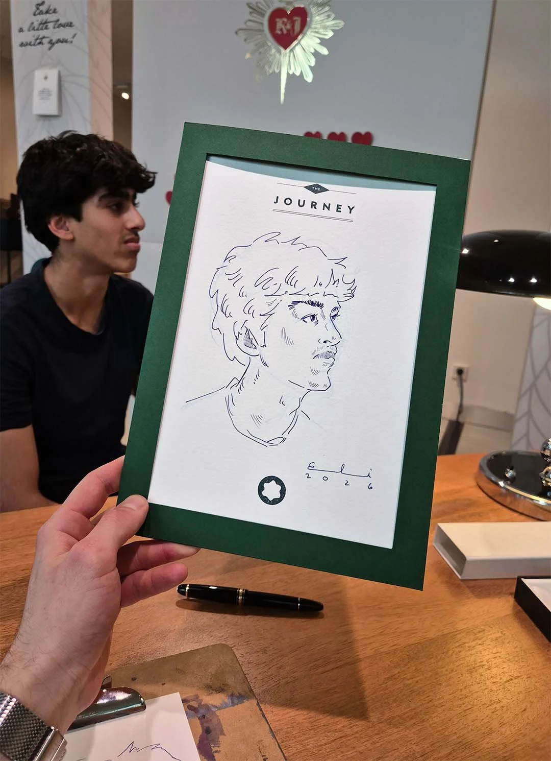 Hand hält einen Bilderrahmen mit einer handgezeichneten Porträtzeichnung eines Mannes. Im Hintergrund ein junger Mann mit schwarzen Haaren, ein Tisch mit Stift und anderem Zubehör.