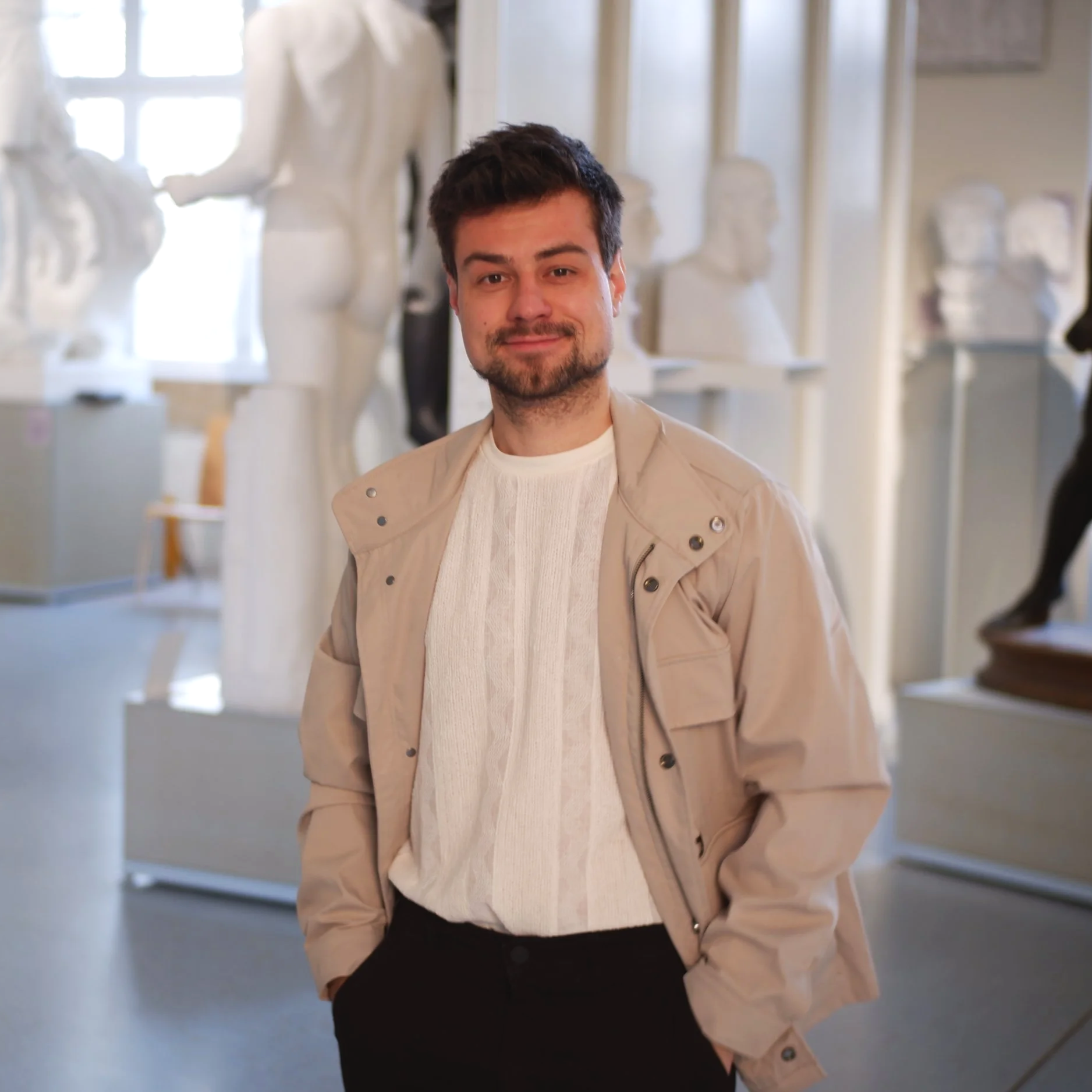 A young man with dark hair and a beard, wearing a beige jacket and white sweater, standing in an art gallery surrounded by sculptures.
