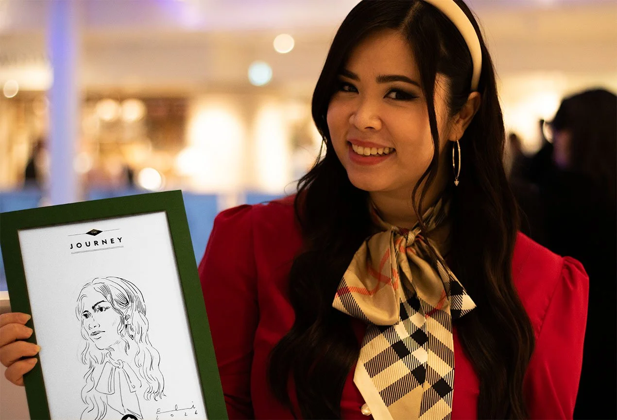 Eine lachende Frau mit dunklen Haaren, rotem Kleid, Headband und Schal, die ein gerahmtes Karikaturbild hält, im Hintergrund eine gemütliche, helle Atmosphäre.