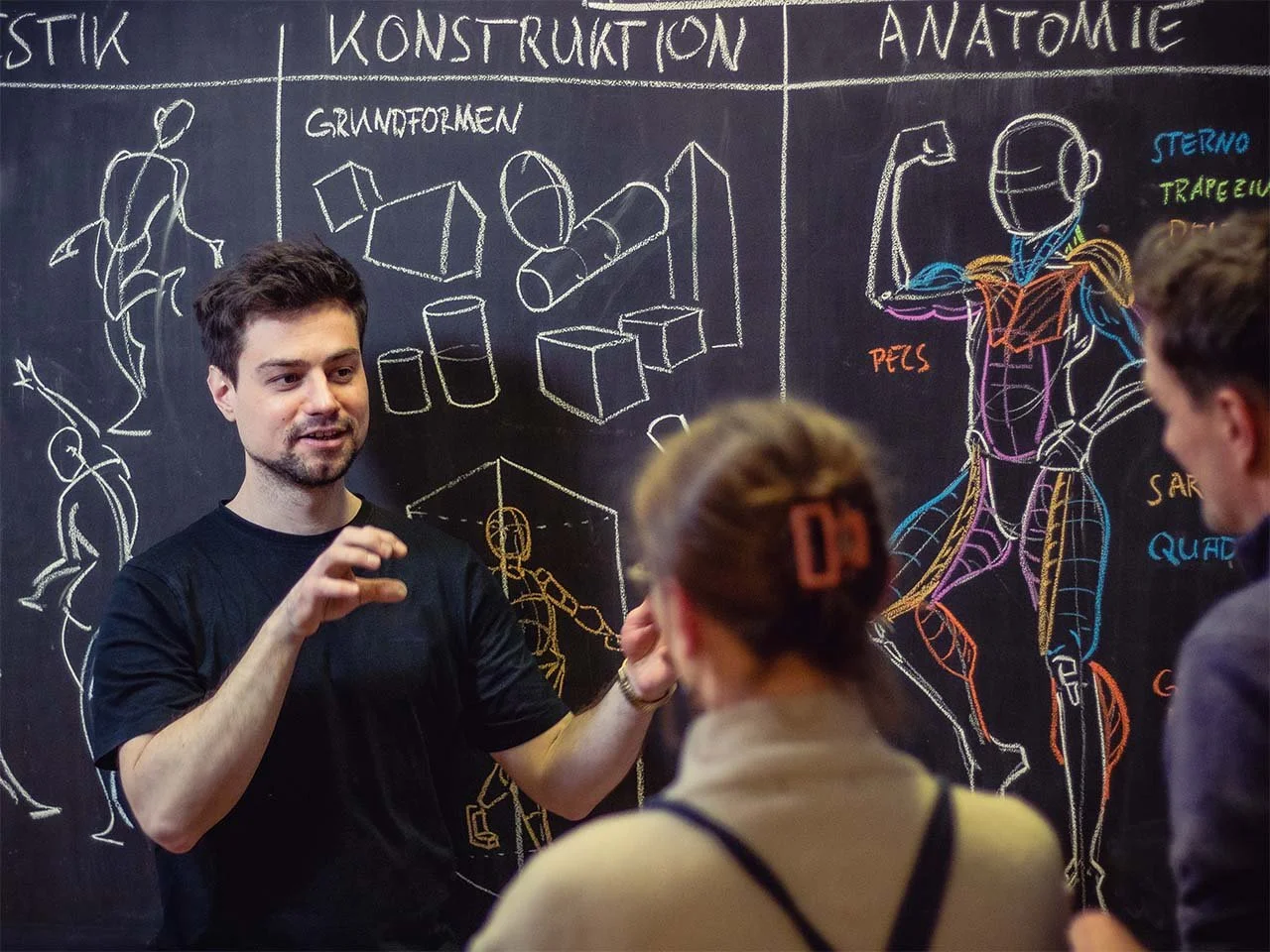 Ein Mann erklärt einer Gruppe von Menschen Anatomie-und Konstruktionszeichnungen an einer Tafel, die auch Skizzen menschlicher Muskulatur und Knochen zeigt.