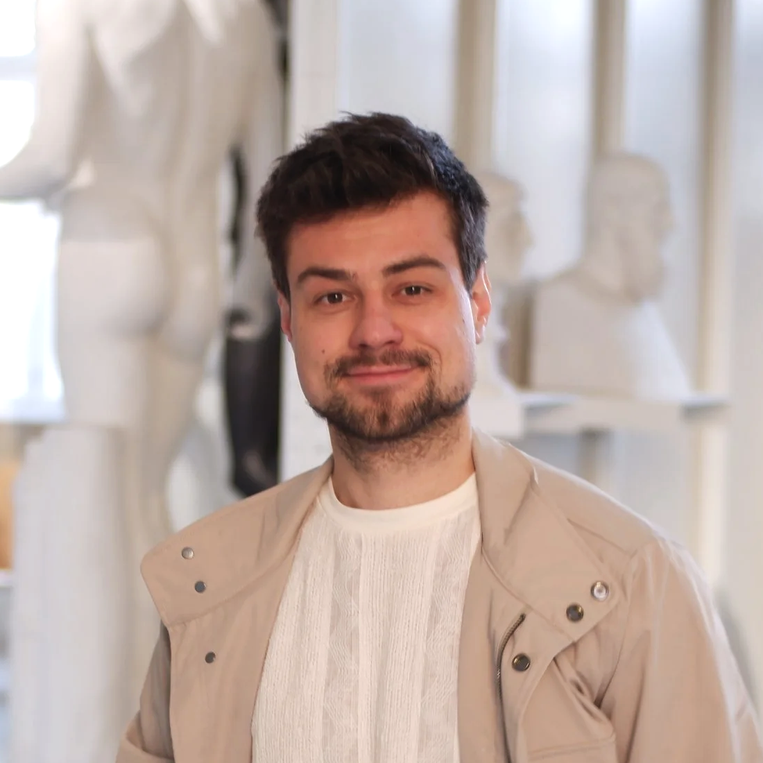 Eli Schneider wearing a beige jacket over a white shirt, standing in front of sculpture busts and artwork in a museum or gallery.