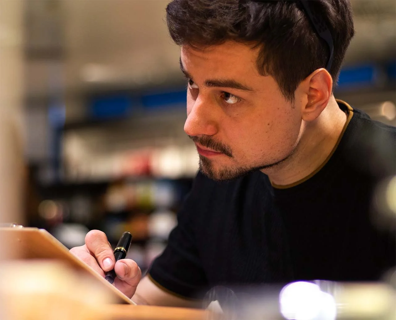 Junger Mann arbeitet konzentriert im Restaurant oder Café, schreibt oder zeichnet auf einem Notizblock, trägt schwarze Kleidung, im Hintergrund Regale mit verschiedenen Gegenständen.