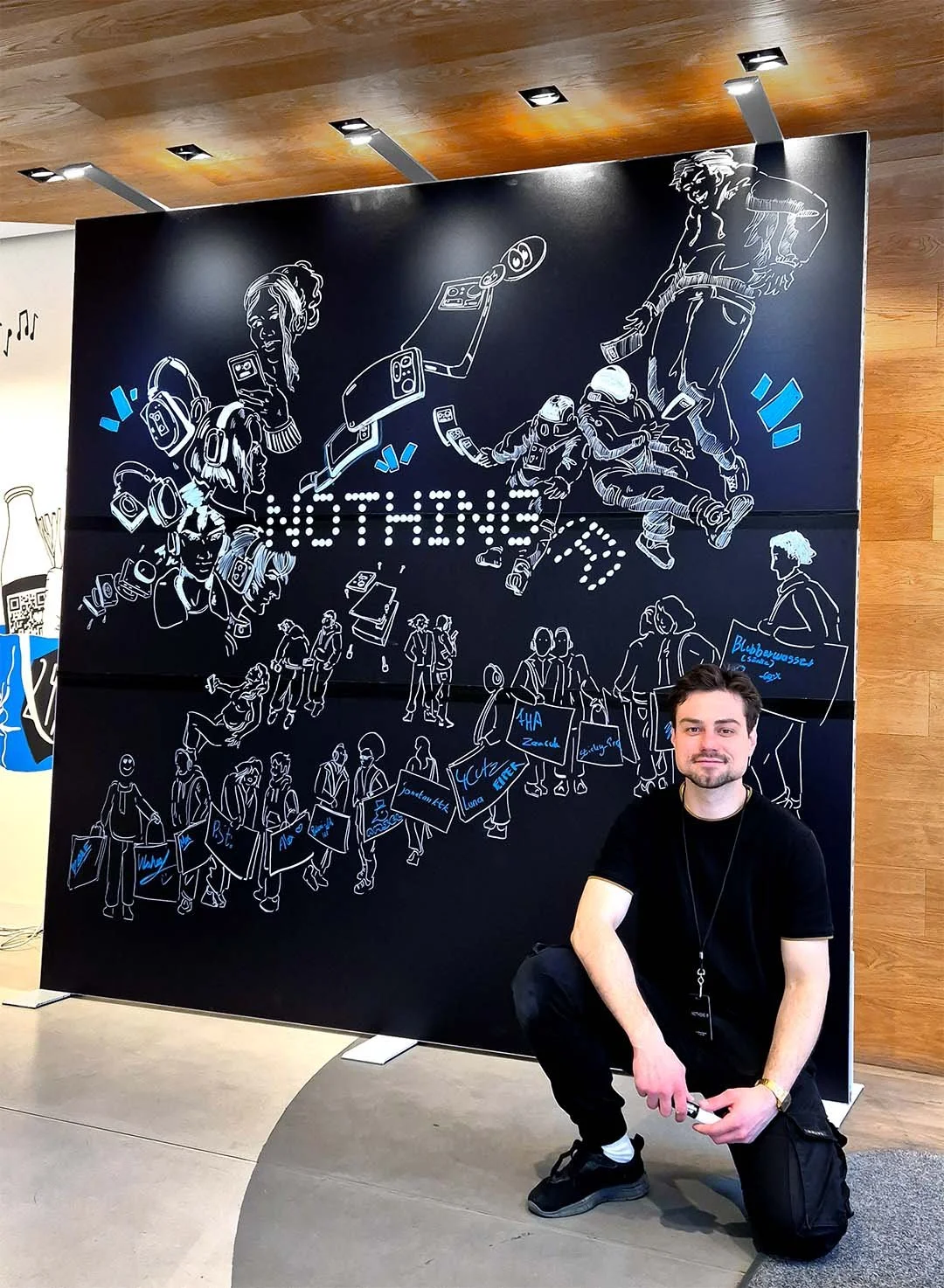 Ein junger Mann posiert vor einer großen schwarzen Wand mit weißen und blauen Linienzeichnungen, die Menschen, Technologie und das Wort 'OTHER' darstellen.