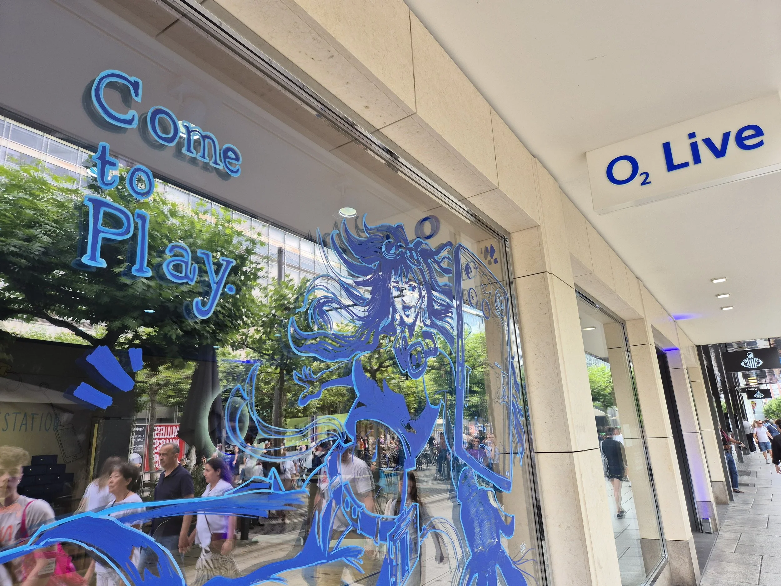 Store window with illustration of a girl with long flowing hair and a nothing smartphone, and the text 'come to play.' The store sign reads 'O₂ Live.' Reflections of people and trees are visible in the glass.