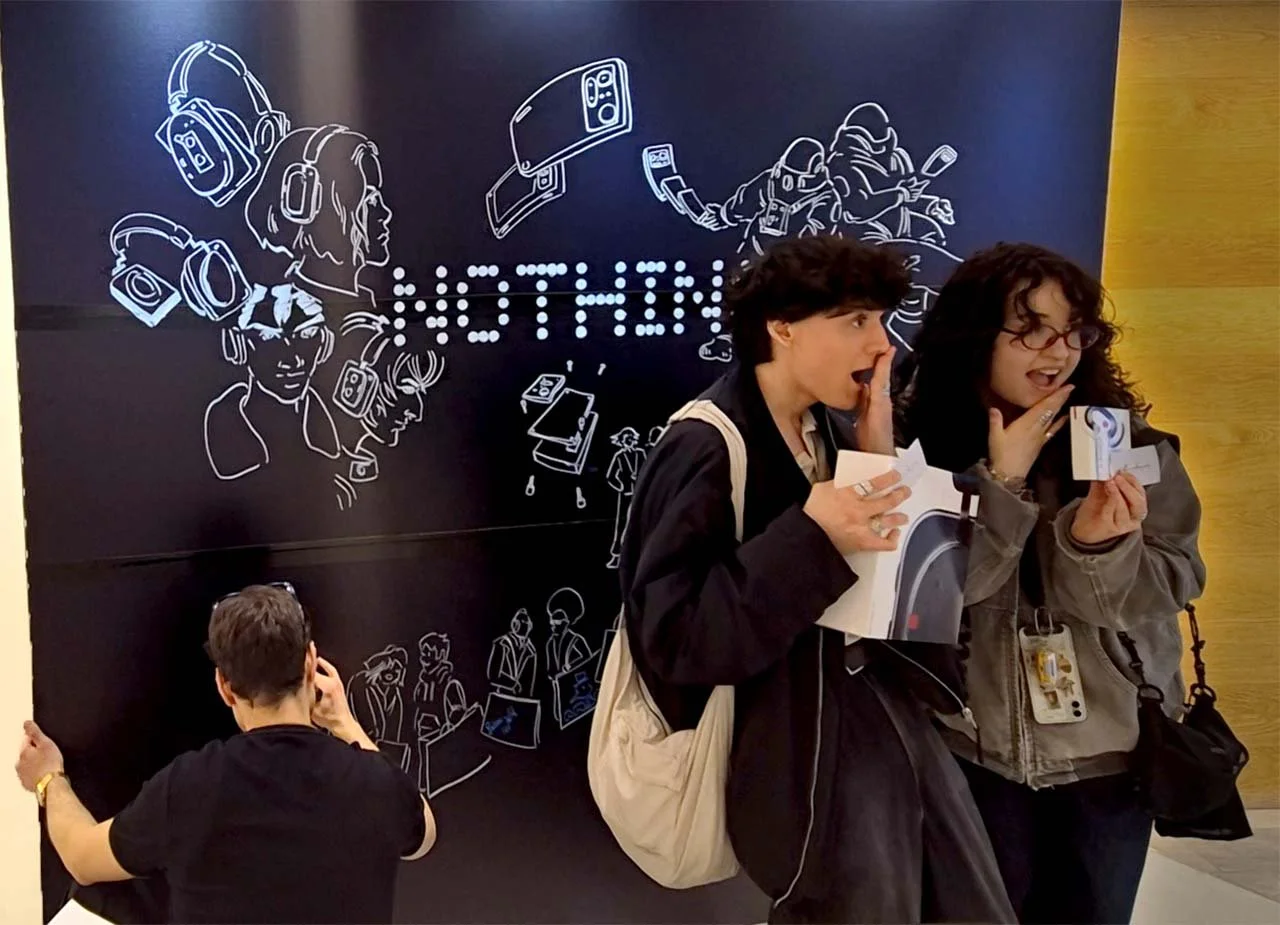 Zwei junge Frauen kommentieren überrascht während einer Veranstaltung, eine hält eine Eintrittskarte, im Hintergrund eine Wand mit Illustrationen von Menschen mit Kopfhörern, Kameras und technischen Geräten.