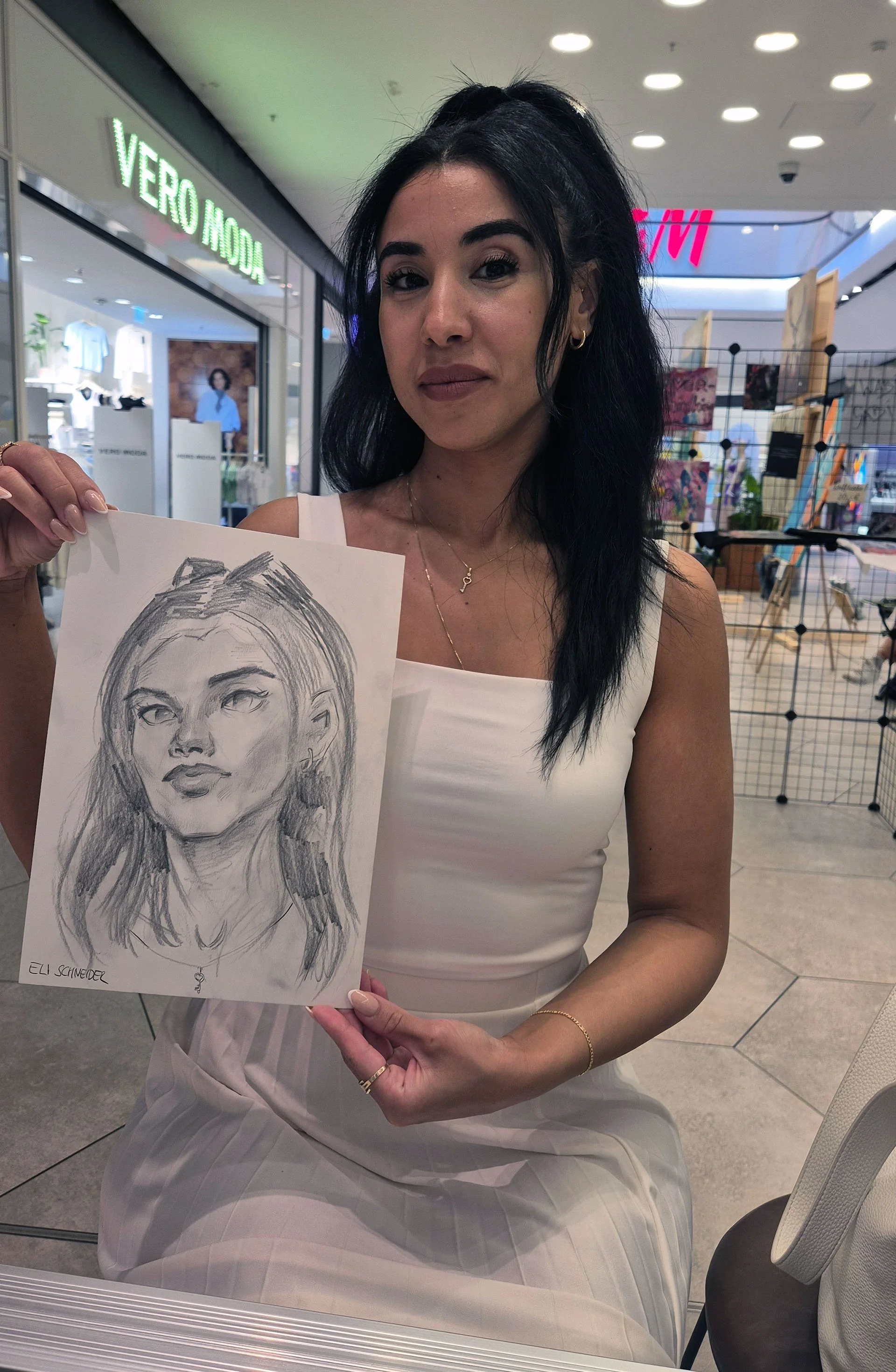 A woman with dark hair, wearing a white dress, sitting indoors and holding a sketch of a woman's face, which she shows to the camera.