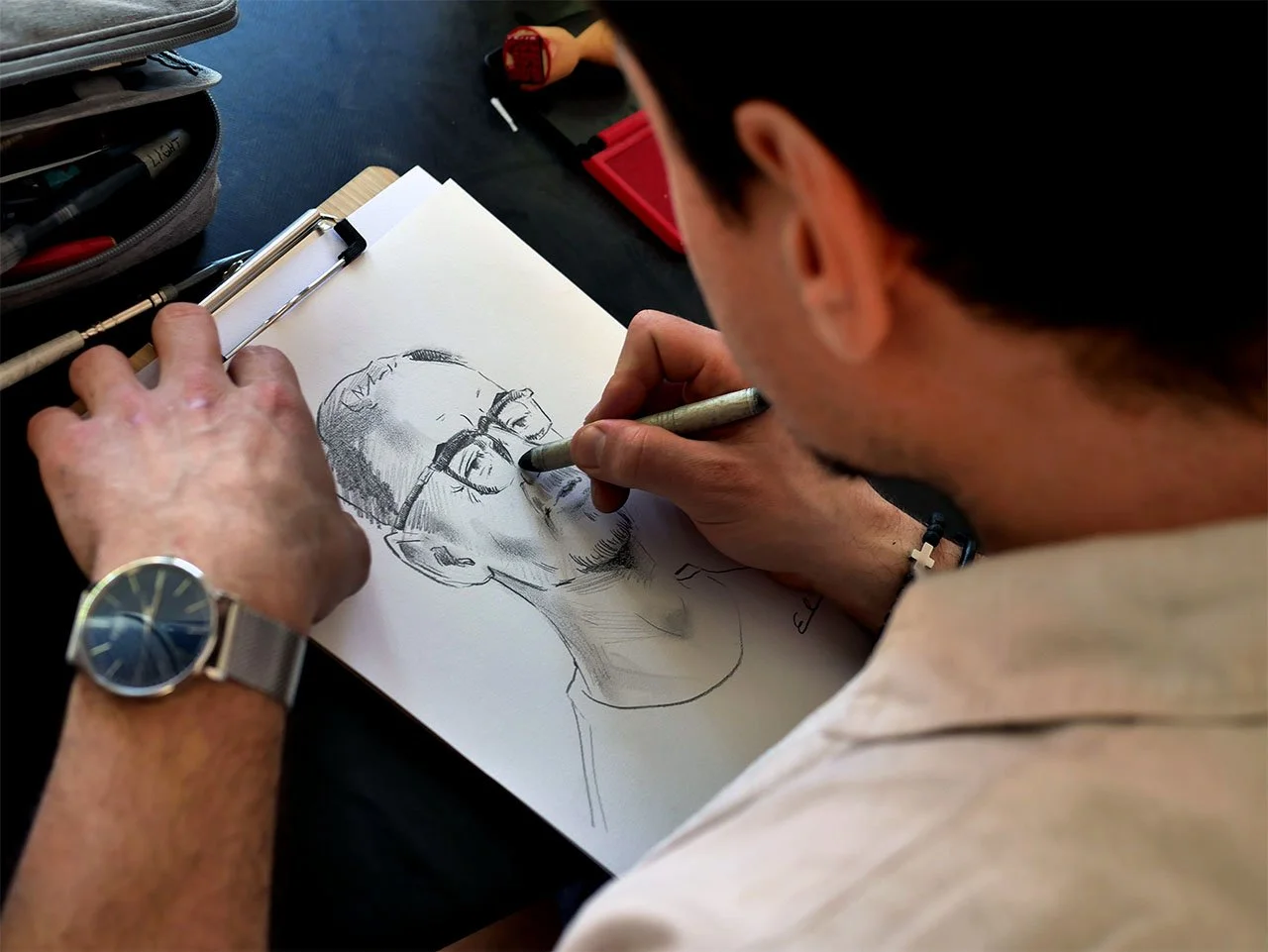 Ein Mann zeichnet eine Skizze eines Mannes mit Brille auf einem Blatt Papier. Er benutzt einen Stift und trägt eine Armbanduhr.