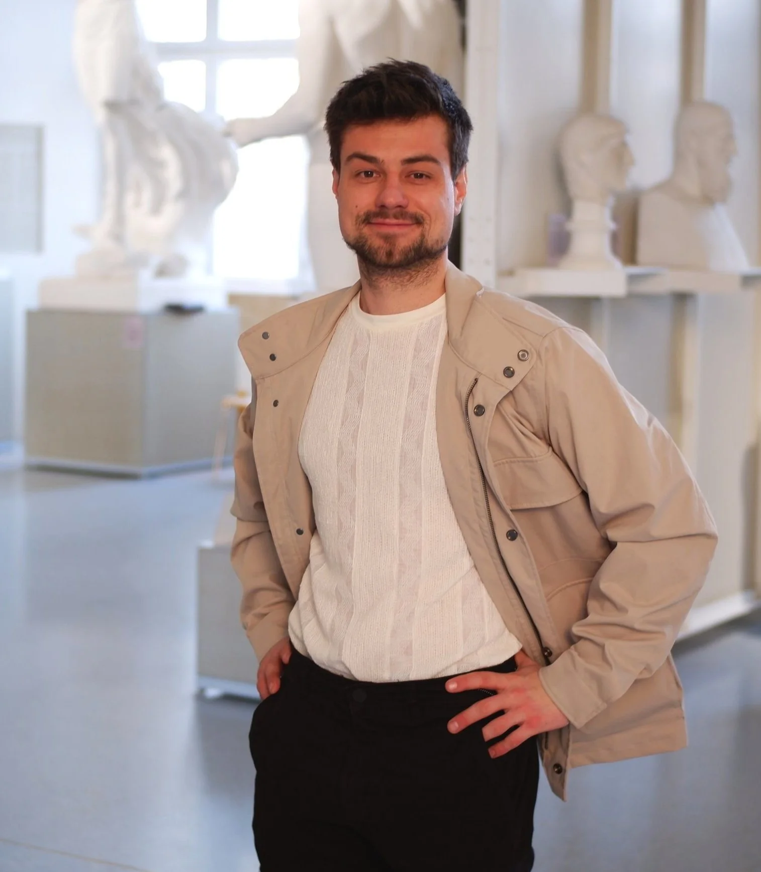 Man in a beige jacket and white shirt standing in an art gallery with white sculptures and busts in the background.
