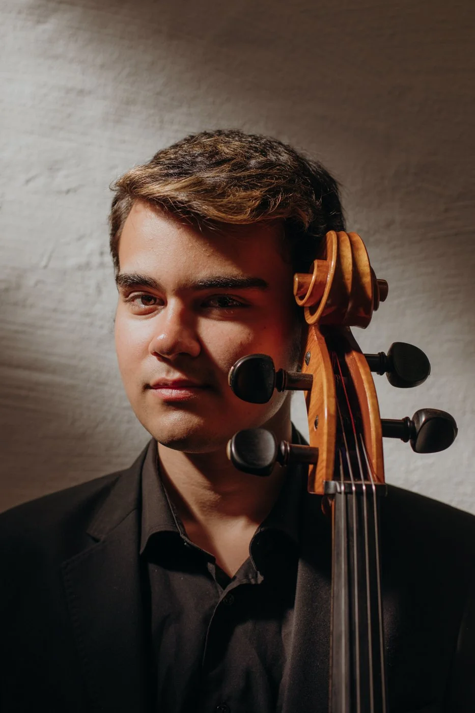 Headshot of a young man in a black shirt, having the pegbox of a cello next to his face