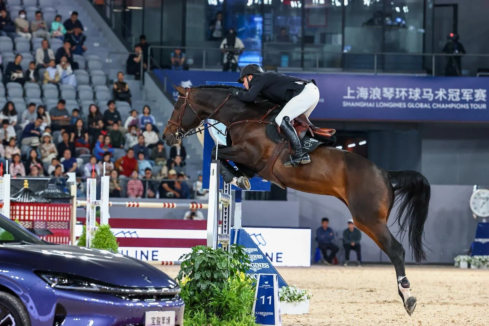 A rider in black and white attire jumping with a brown horse over an obstacle during an equestrian event at a stadium. Spectators are seated in the stands watching.