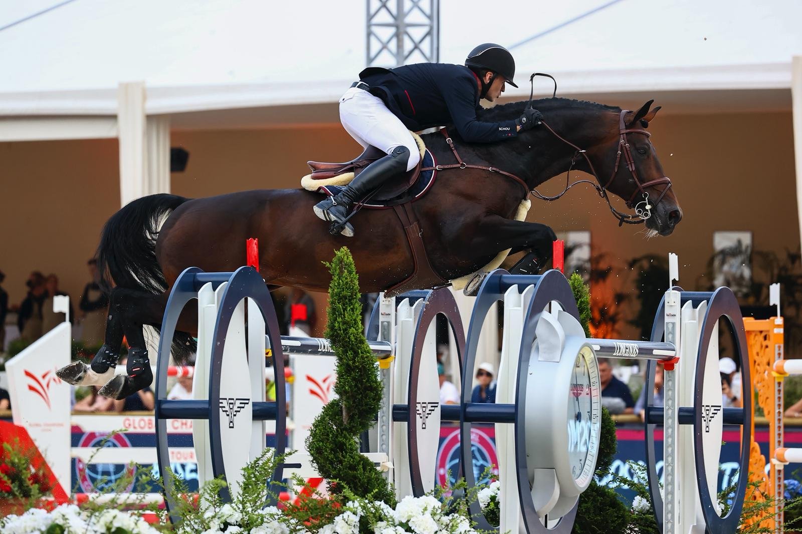 A person in riding gear horseback jumps over an obstacle with a clock face design in an equestrian show jumping competition.