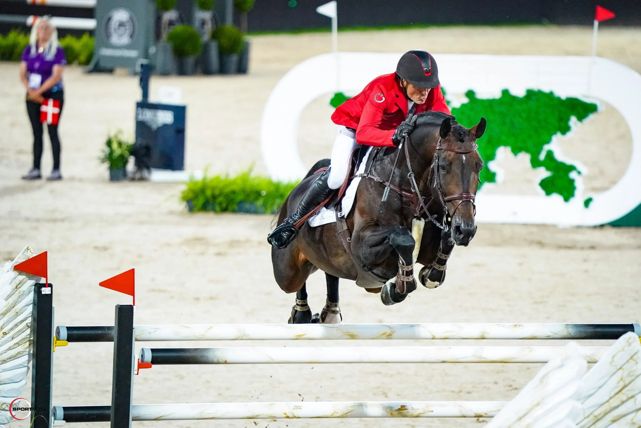 A rider in a red jacket and black helmet jumping a horse over an obstacle in an equestrian show jumping event.