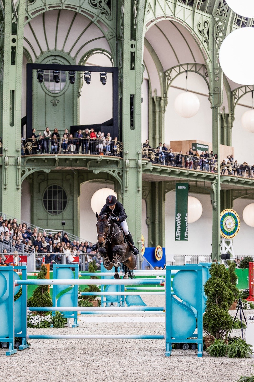 A horse and rider competing in an indoor show jumping event, with spectators watching from balconies and stands in a large, ornate, green-domed arena.