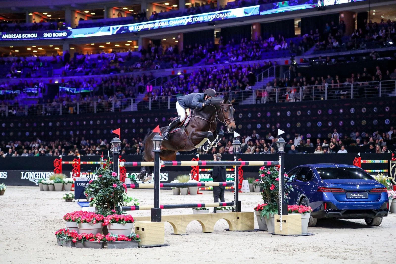 A man riding a horse in an indoor equestrian arena, jumping over an obstacle that includes a car, with a large audience watching from the stands.