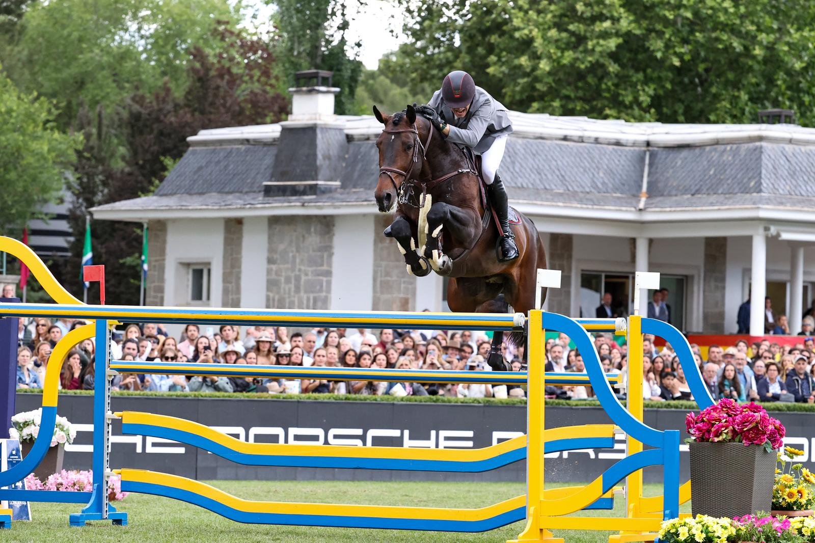 A jockey on a brown horse jumping over a colorful obstacle during a show jumping competition, with an audience watching in the background.