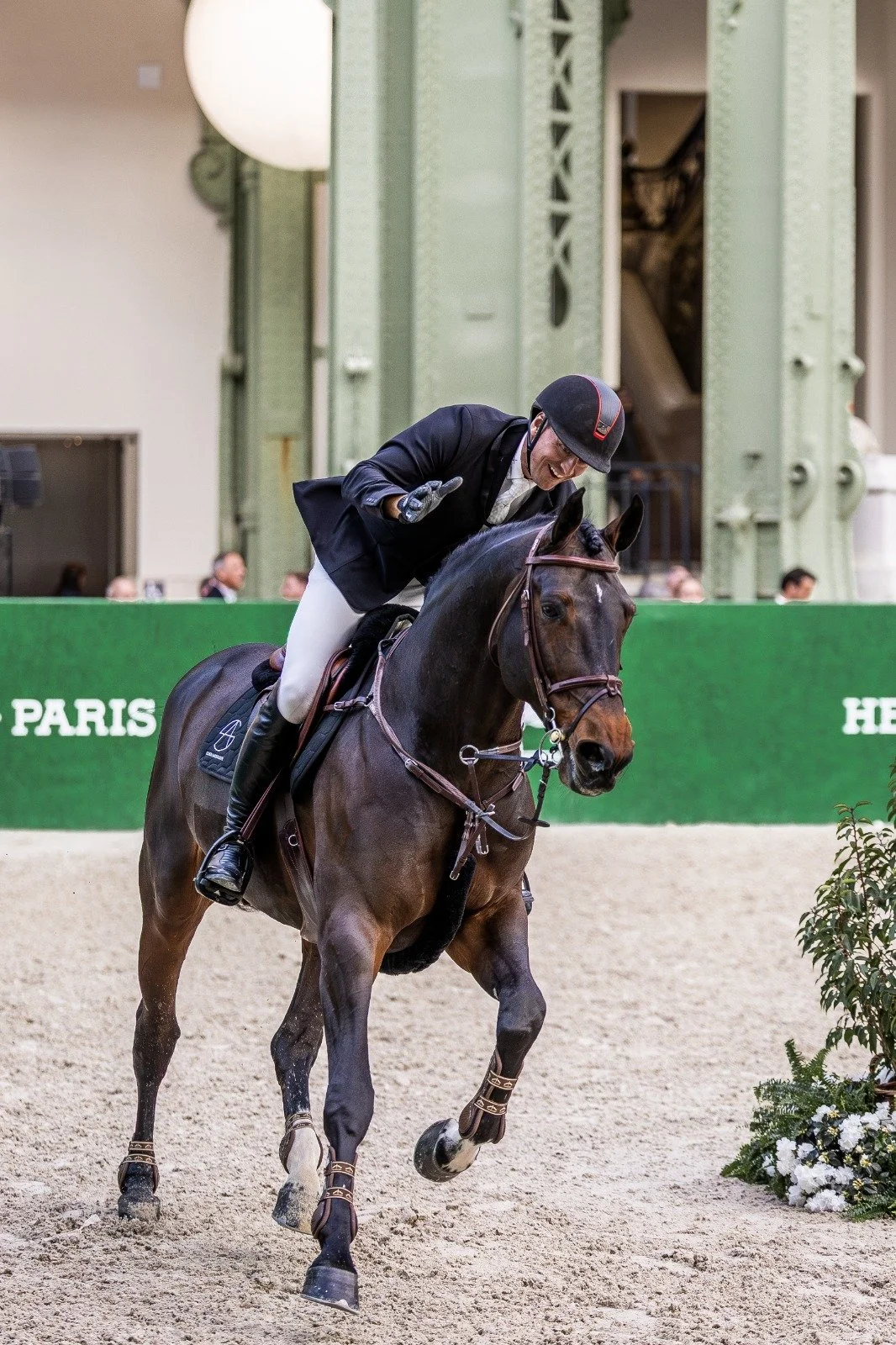 A male equestrian rider wearing a black jacket, white pants, a helmet, and gloves, smiles and gives a thumbs up while riding a brown horse in an indoor arena.