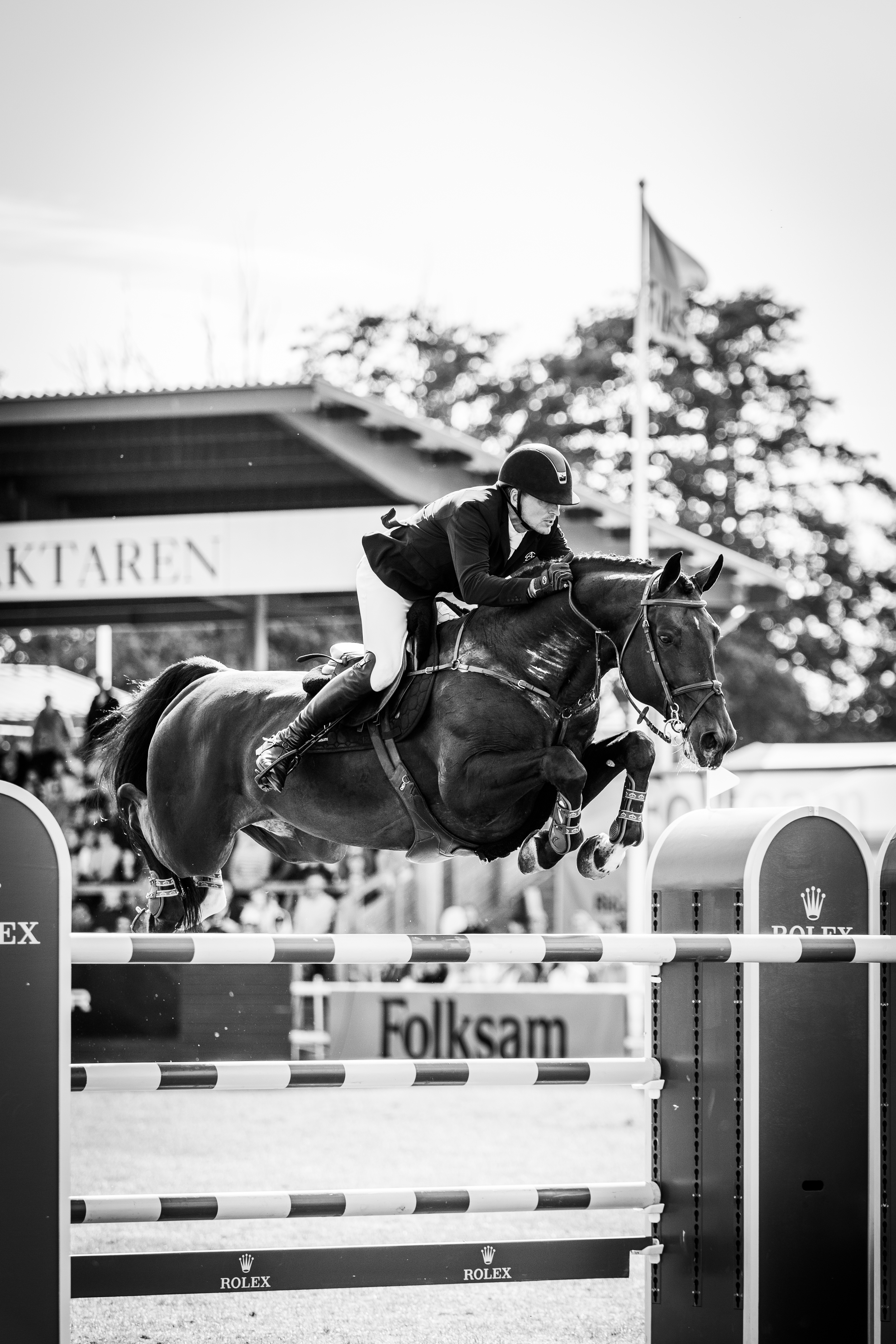Black and white photo of a male equestrian riding a horse over a show jumping obstacle at a competition, with spectators and banners in the background.