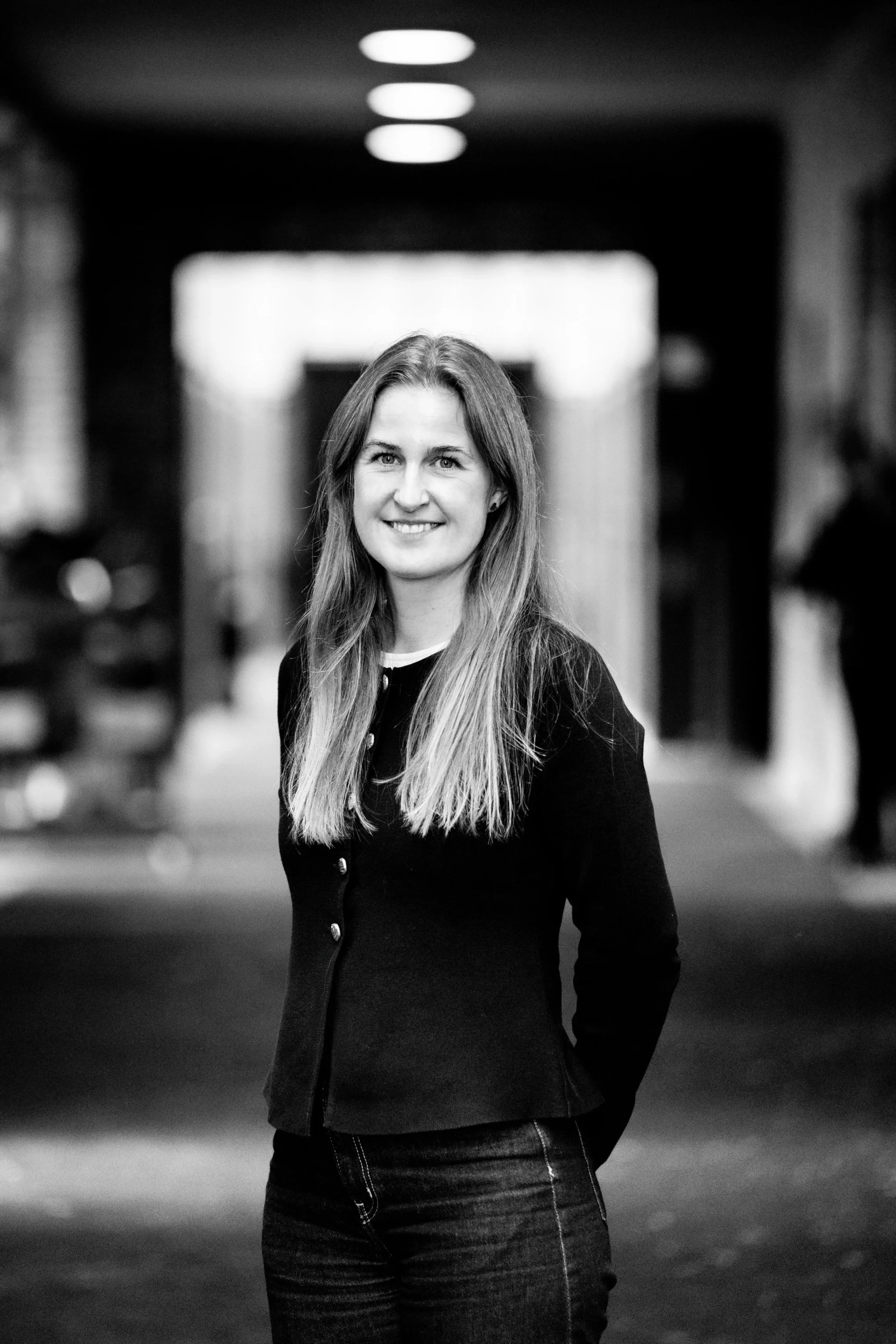 Black and white photo of a woman standing in a hallway inside a building, smiling and looking at the camera.