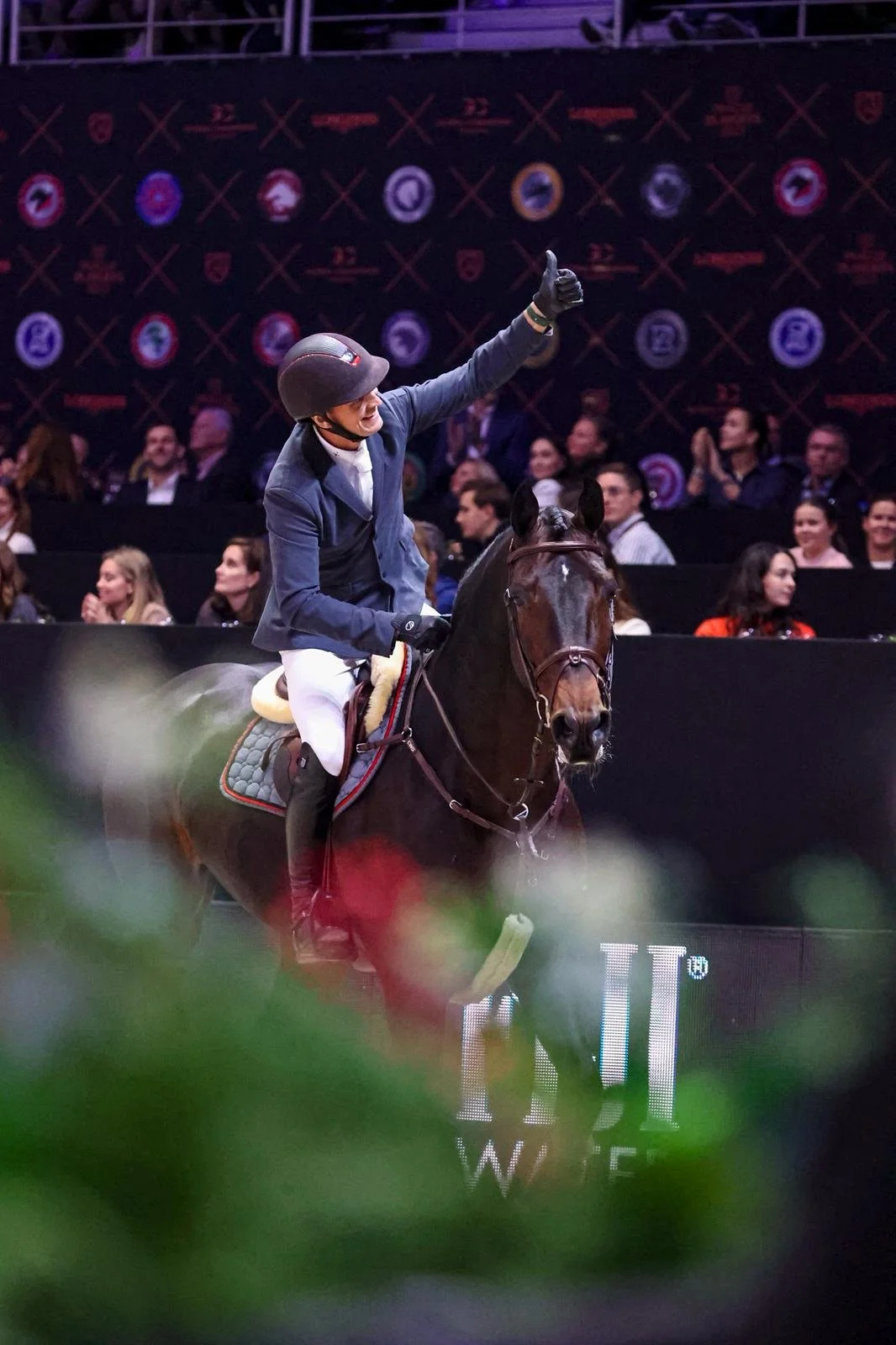 A person riding a horse at an indoor equestrian event, giving a thumbs-up sign with the crowd clapping in the background.