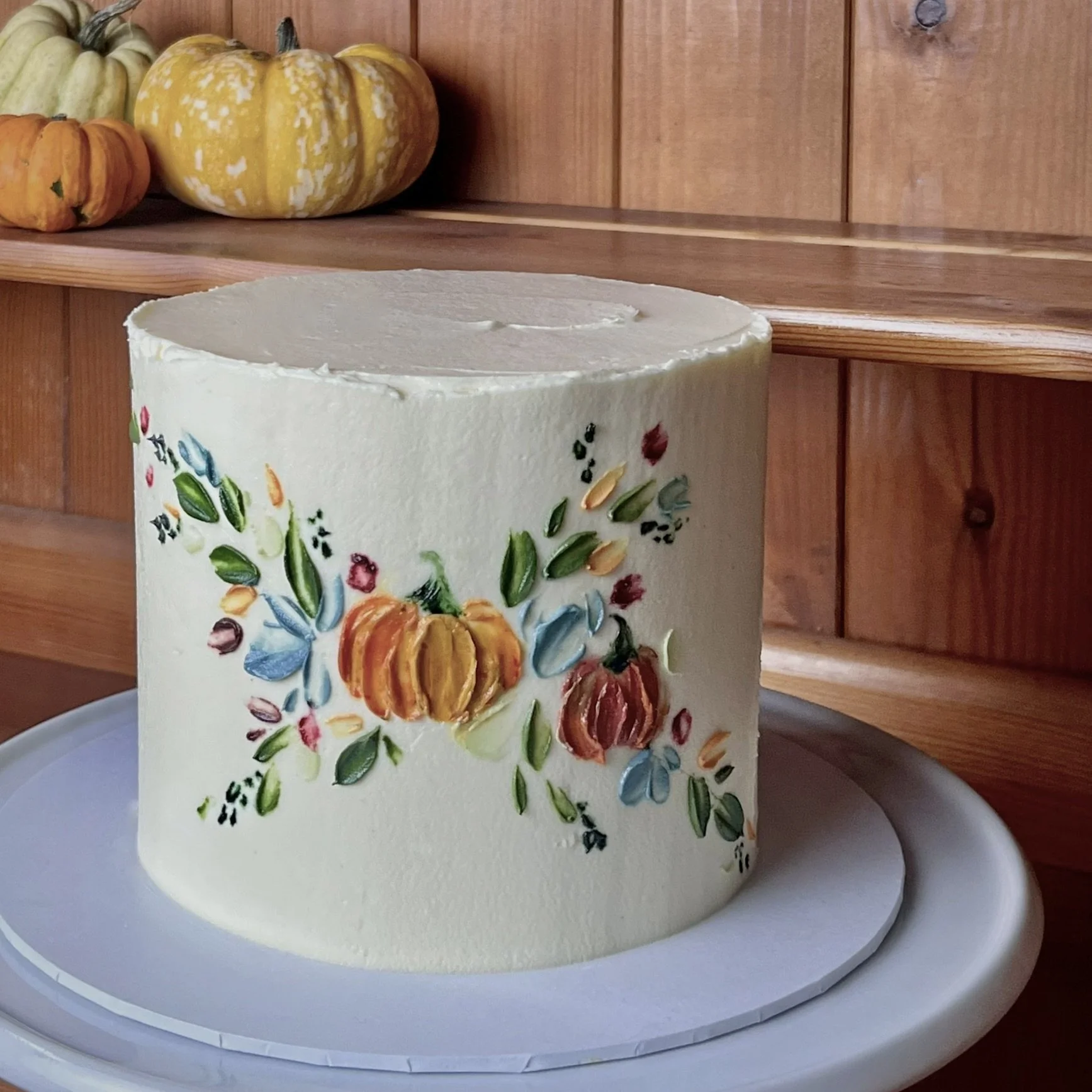 A white frosted cake decorated with hand painted colourful autumn-themed floral designs including pumpkins and leaves.