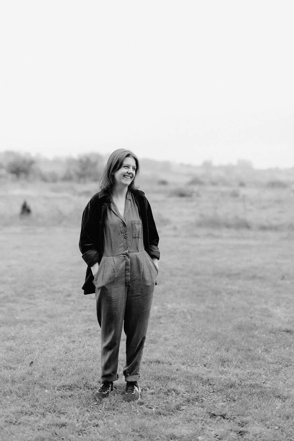 A wedding cake designer stands outdoors on a grassy field looking for inspiration, smiling and looking to her left, with trees and distant landscape in the background.