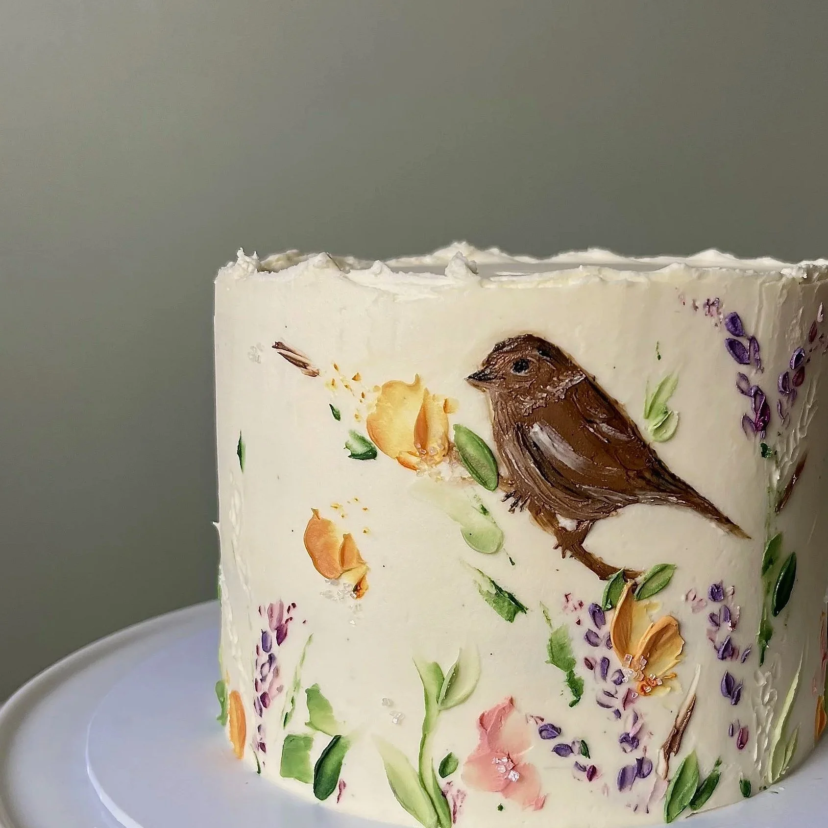 A cream coloured cake with colourful wildflowers, petals and folliage with a garden bird resting on a branch