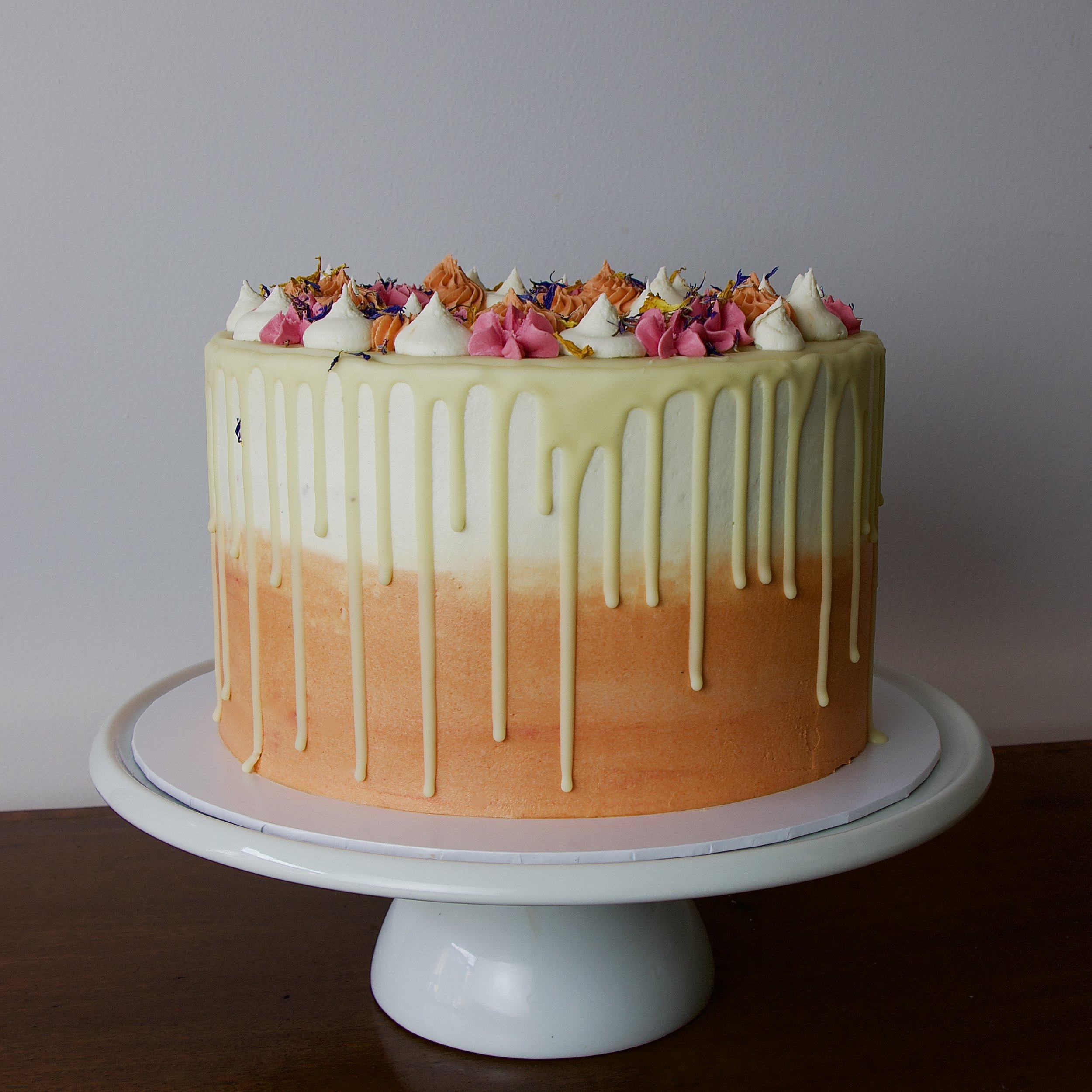 A round cake with an orange ombré bottom tier and a white top tier, decorated with a white chocolate drizzle and colorful flowers on top, placed on a white cake stand.