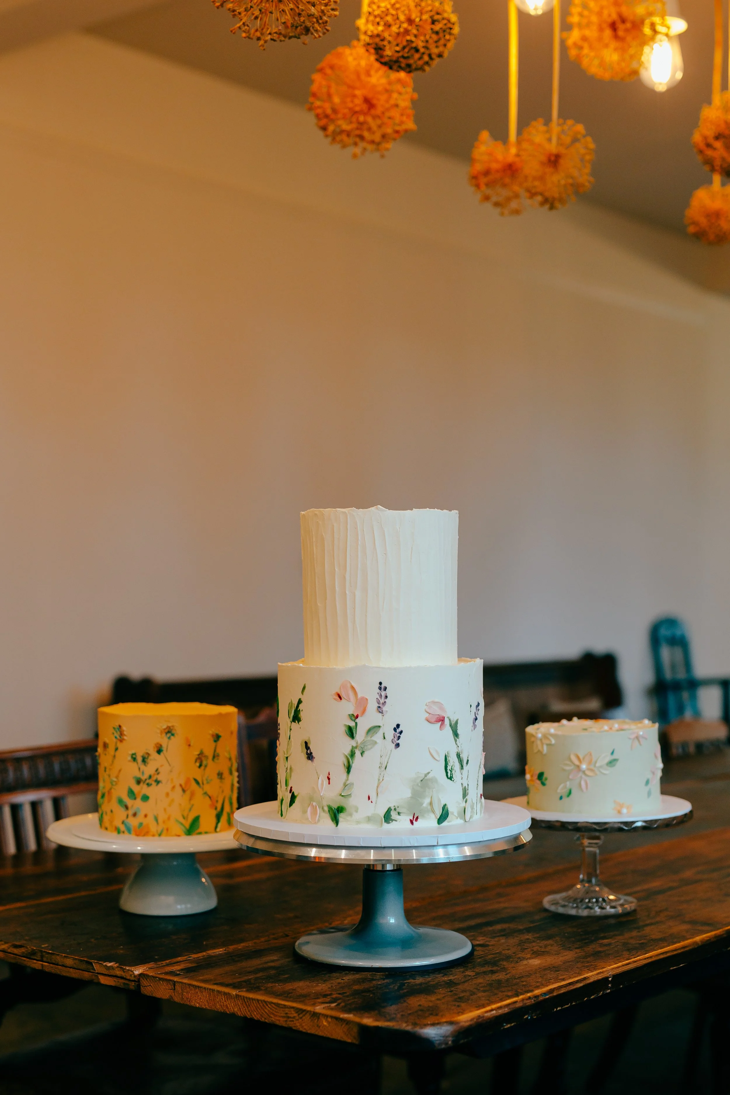 Three wedding cakes lines up next to each other, each with a natural, floral and wildflower design feating a white wedding cake in the centre and colourful wedding cakes either side