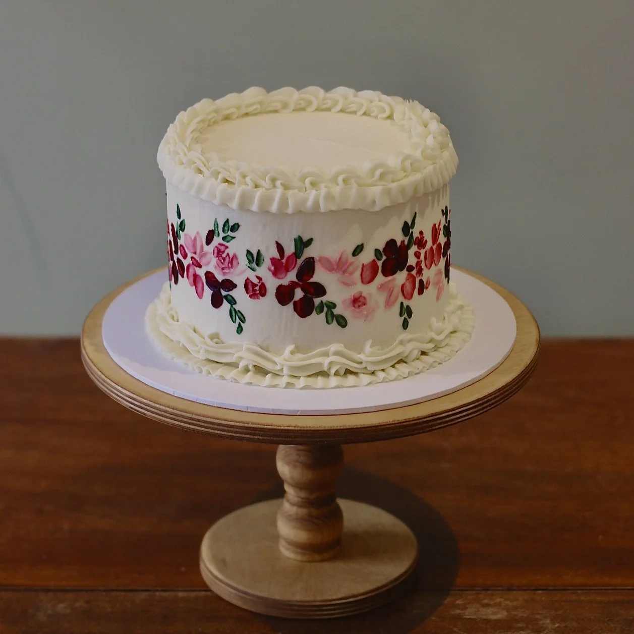 A single tiered wedding cake with vintage piping and a hand-painted floral design around the middle on a wooden cake stand