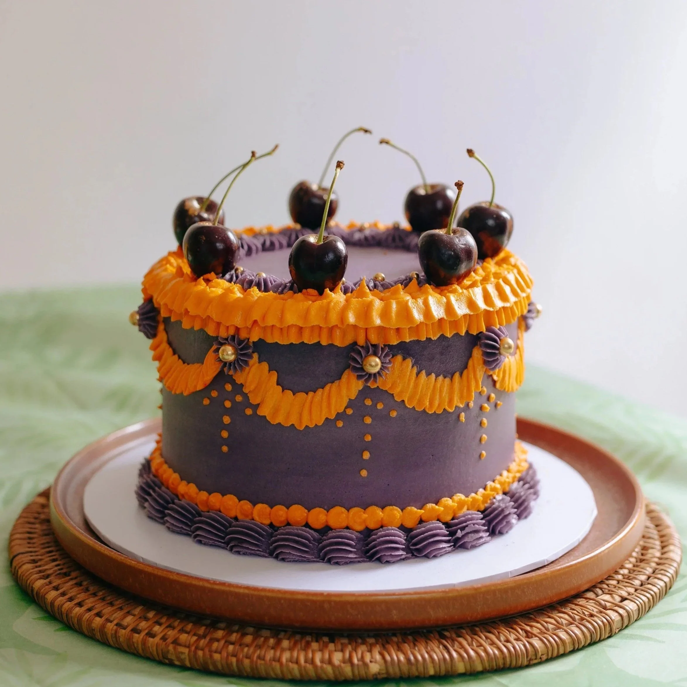 Purple vintage birthday cake decorated with orange and purple icing, topped with cherries and gold accents, placed on a wooden and white cake board.