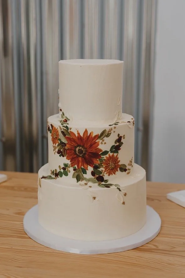 A three tiered wedding cake with a whimsical floral painted design on the centre in burnt oranges and forest greens