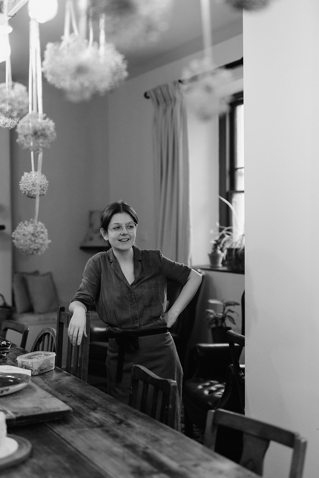 A cake designer in a dining room, smiling, with arms resting on chairs, decorated with hanging natural flower arrangements.