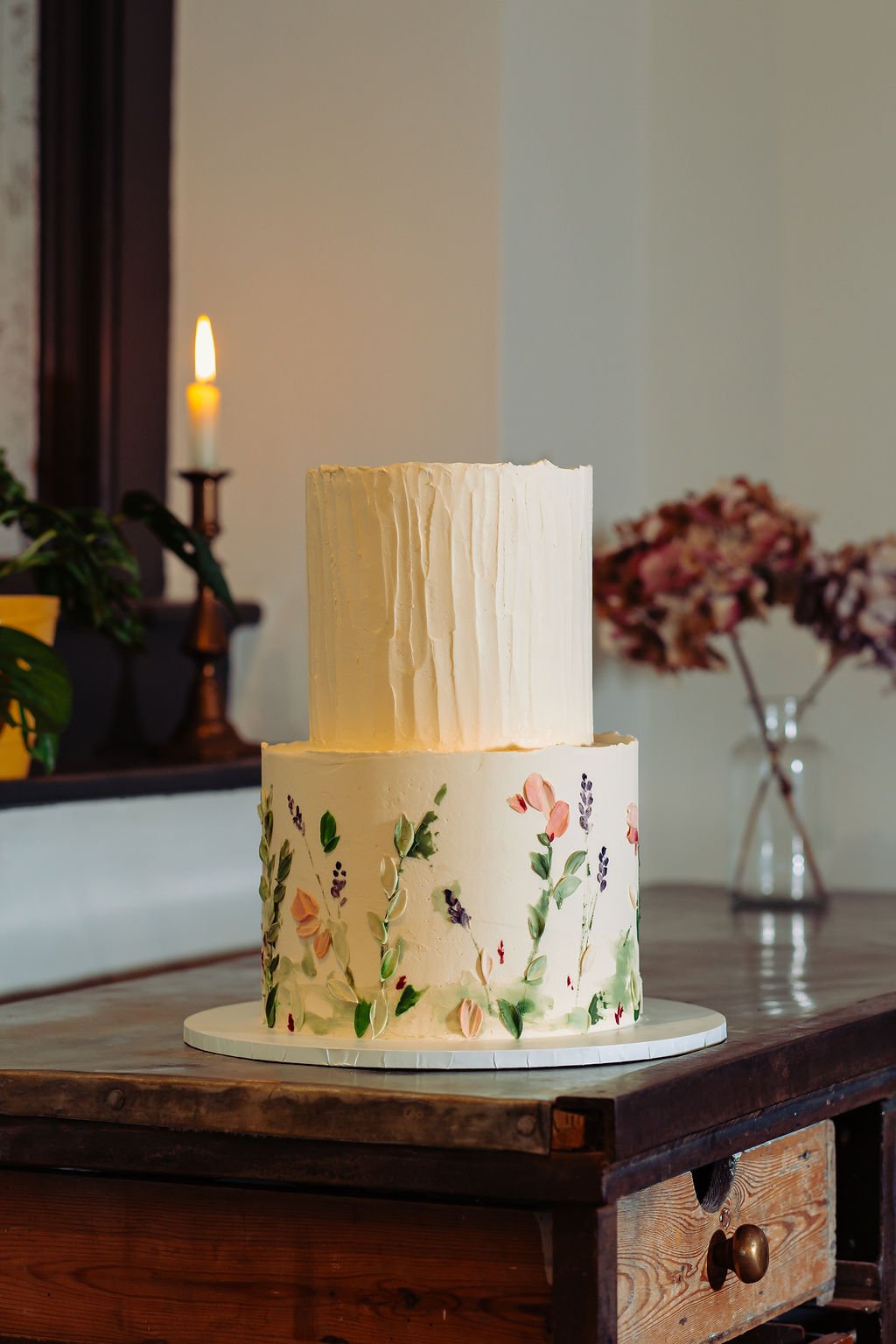 A white two tier vegan wedding cake with a wildflower meadow theme on the bottom tier and rustic buttercream icing on the top tier