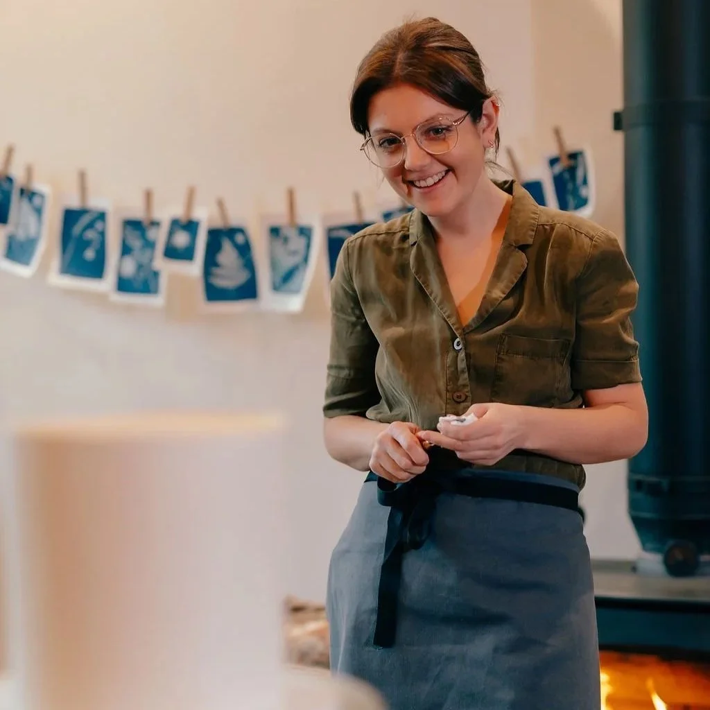 A smiling woman with short brown hair and glasses, wearing a green shirt and gray apron, standing indoors with photos hanging on a string in the background.